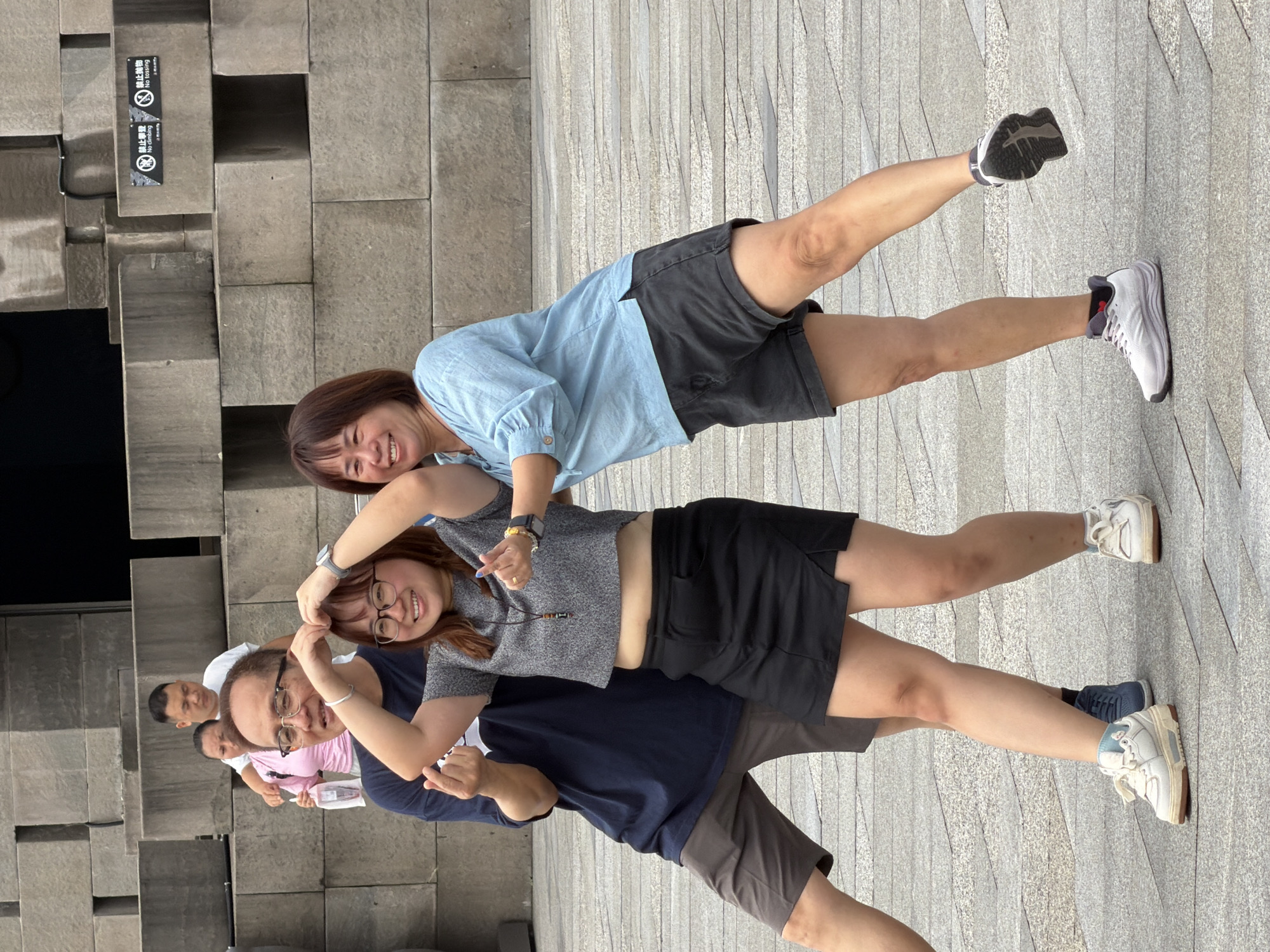 In Yuzhong District, China, three adults pose for a photo in front of a textured stone wall. A man wearing glasses and a dark blue t-shirt stands on the left, with his left arm around a woman in the center. The woman, wearing a gray crop top and a black skirt, smiles and makes a heart shape with her fingers above her head. To the right, another woman in a light blue shirt and dark shorts smiles and gestures with her hands. The ground is paved with large, square tiles.