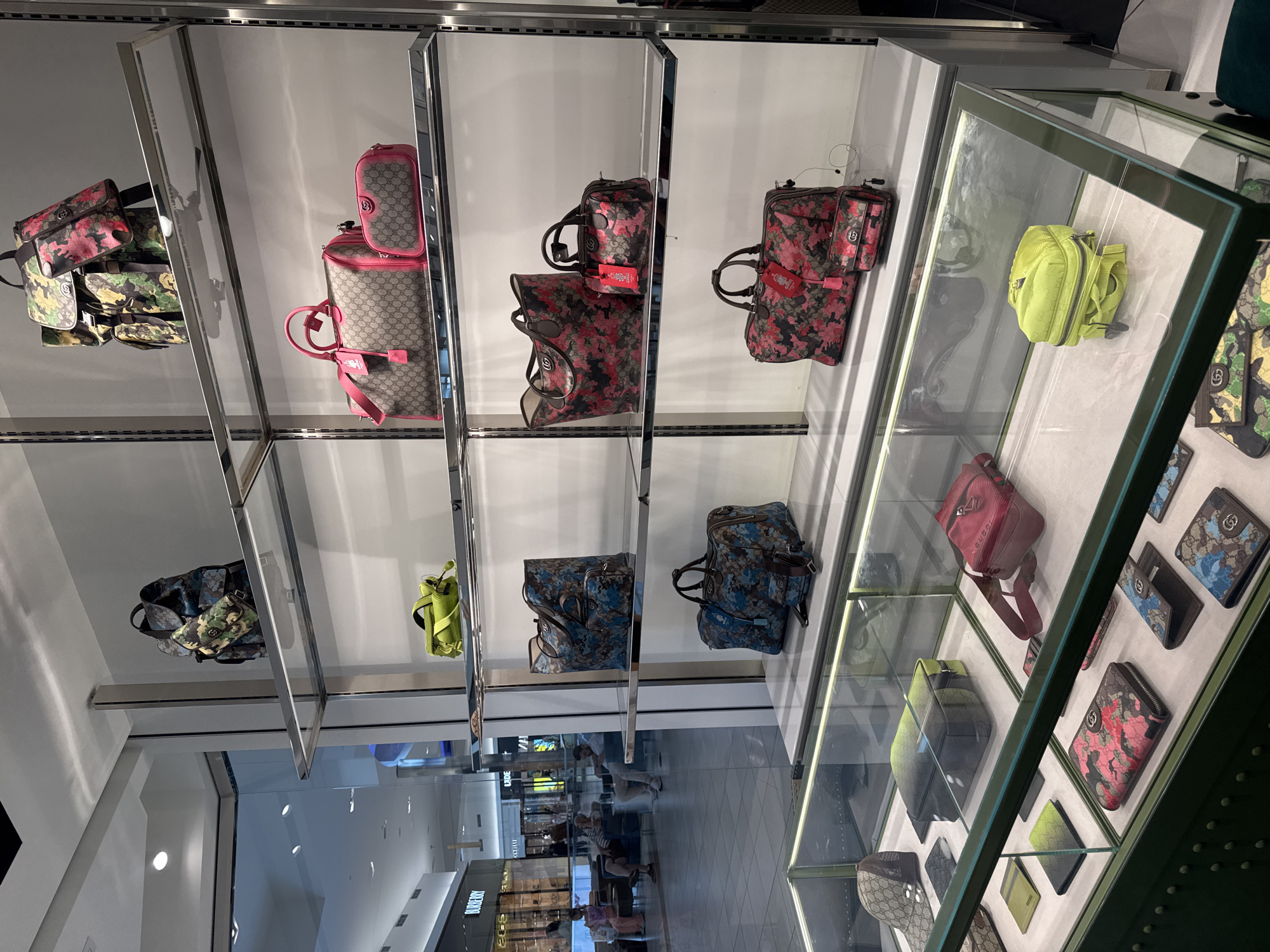 The image displays an indoor retail setting, likely a luxury fashion boutique or a dedicated section within a department store, located in Rosemont, United States of America. The brightly lit scene features a modern display system with glass shelves and green-trimmed glass display cases showcasing various handbags and wallets.

The primary subjects are numerous luxury accessories, predominantly from Gucci, identified by the distinctive "GG" monogram pattern and brand tags. The collection includes handbags, duffle bags, crossbody bags, and wallets. Many items feature a camouflage-inspired pattern in different color schemes, notably pink/red, blue, and green/brown. Some bags blend the camouflage print with the classic "GG" monogram, often accented with vibrant pink details. Specific items visible include a large pink monogram tote with a matching small pouch, several duffle-style bags in pink/red and blue camouflage, a lime green crossbody bag, and various wallets matching the different camouflage patterns.

In the background, through reflections on the polished floor and glass panels, several out-of-focus people can be seen. These individuals appear to be shoppers or mall patrons, some walking and one seated, suggesting a typical shopping environment. The activity depicted is browsing or purchasing high-end fashion accessories.

Notable details include the crisp, clean aesthetic of the display, emphasizing the products. Faint reflections of other store signs, such as "BURBERRY," are visible in the background, confirming the location as part of a larger shopping mall. The time of day is not explicitly indicated but appears to be daytime due to the bright interior illumination. Visible text includes the "GUCCI" brand name on product tags and the ubiquitous "GG" logo on the merchandise.