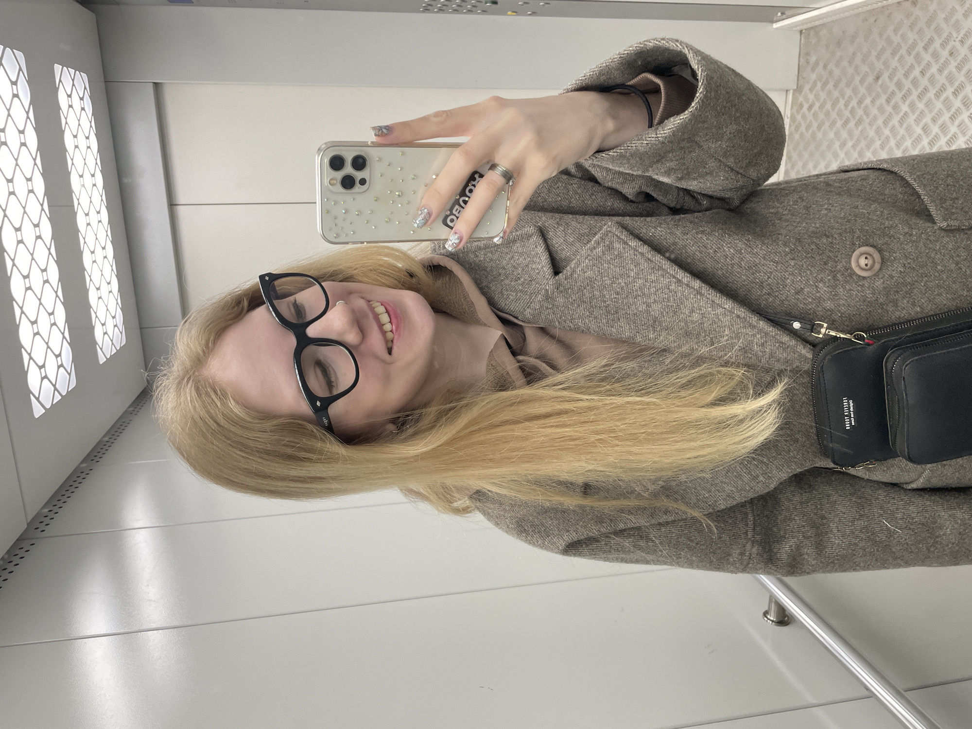 A woman is taking a selfie in an elevator. She has long blonde hair and is wearing black glasses and a tan coat. She is holding a phone with a decorated case in her right hand. Her nails are also decorated. A black purse is slung across her body. The elevator has white walls and a metal handrail. The location is likely in Izhevsk, Russia, based on the user's prompt.