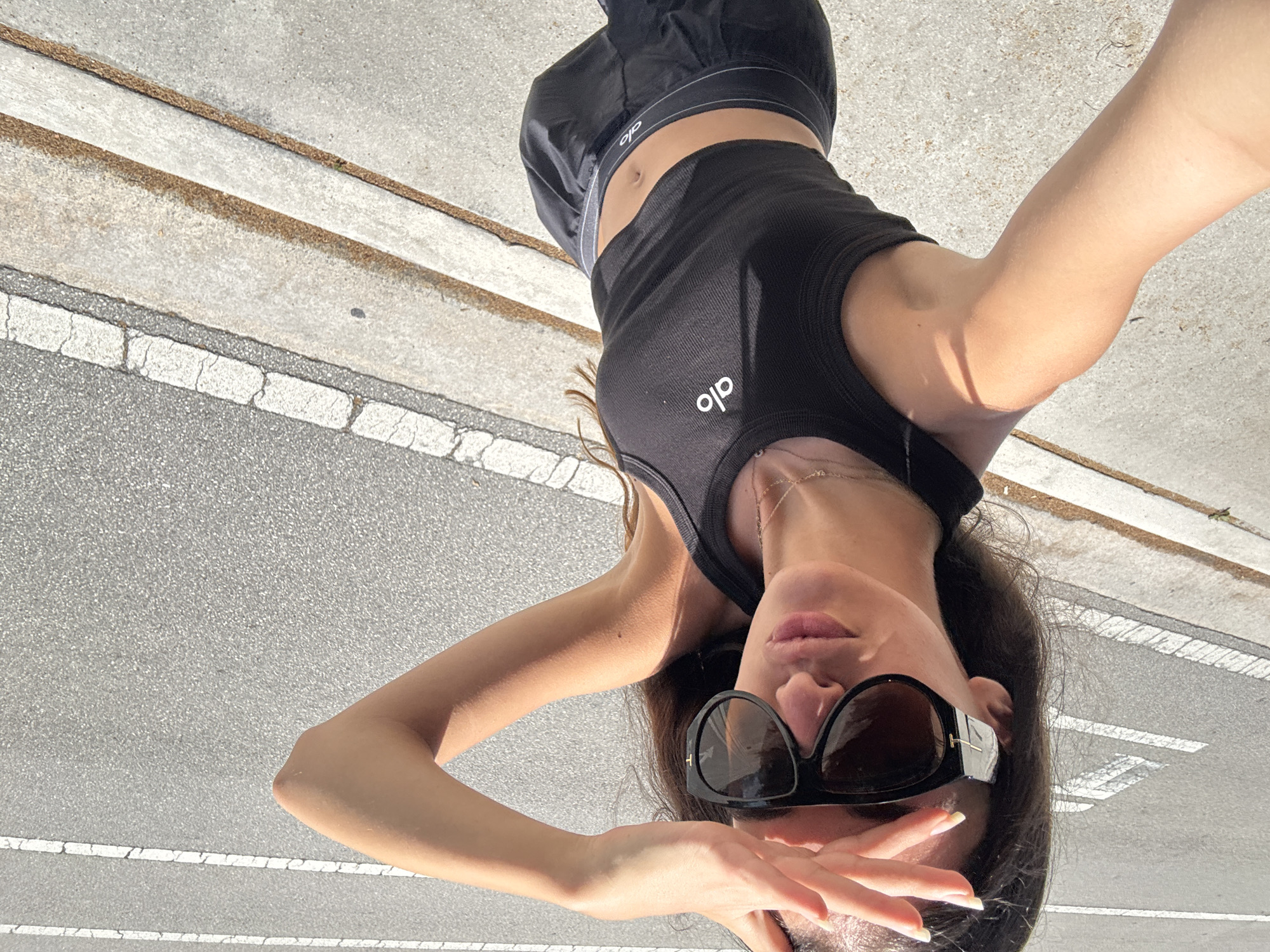 A young woman with long, dark hair and fair skin is posing for a selfie outdoors. She is wearing oversized black sunglasses with silver accents, a black cropped tank top with a white logo, and black shorts with a gray waistband that also features a white logo. Her right hand is raised to shield her eyes from the sun. She is standing on a sidewalk next to a road with white markings, likely in a city setting. The background consists of asphalt and concrete. The lighting suggests it is daytime and sunny.