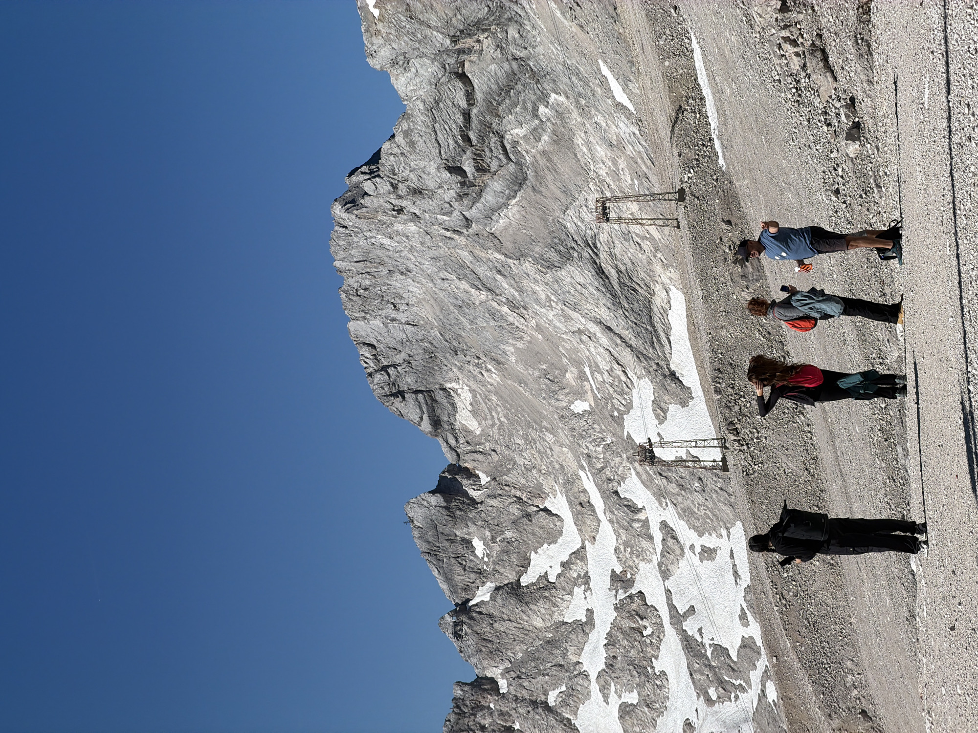 This is a wide shot of a mountainous landscape with a clear blue sky. In the foreground, four people are standing on a gravelly slope. On the left, a person wearing a black hooded jacket and black pants holds a phone in their right hand and has a black backpack. To their right, a person with long brown hair wears a red jacket and black pants and has a red backpack. Next to them, a person with short brown hair wears a blue and gray jacket, blue jeans, and carries a black bag over their shoulder. Finally, on the far right, a person wearing a blue t-shirt, black shorts, and a black baseball cap holds a bottle and gestures with their right hand. In the midground, there are two rusty, metallic towers, possibly part of an old cable car system, with cables extending between them. The background features a large, rugged mountain range with patches of snow scattered across its rocky surface. The image appears to be taken at the Zugspitze, Germany's highest peak, located near Garmisch-Partenkirchen.