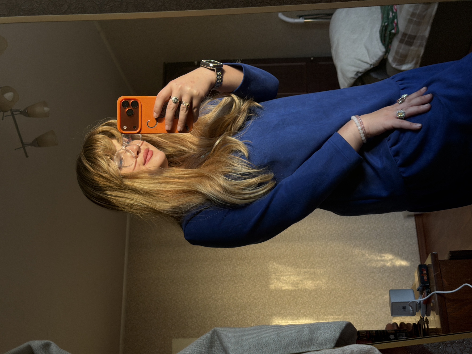 A person is taking a selfie in a mirror. The person has long blonde hair, bangs, and glasses, wearing a blue long-sleeved shirt, multiple rings on both hands, and a watch on their left wrist. They are holding an orange smartphone. The background features a wall, a lamp, a door, and a bed. The location is likely indoors in Naberezhnye Chelny, Russia, given the prompt's context.