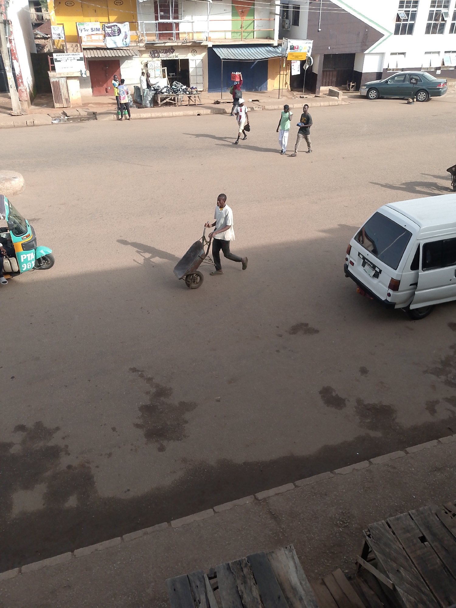 A street scene in Bukuru, Nigeria features a man pushing a wheelbarrow filled with debris. The man is wearing a light-colored shirt and dark pants. He is in motion, walking from left to right across the paved street. To the left of the man, a light blue and green auto-rickshaw is parked, with a person visible in the driver's seat. Further to the left, a green sedan is parked on the side of the road. In the background, buildings line the street, with storefronts and awnings visible. Several other people are walking in the distance, near the buildings. The street surface appears to be asphalt or similar paving material, with some darker patches that could indicate wetness or stains. The overall lighting suggests it is daytime, with shadows cast by the man and the vehicles.