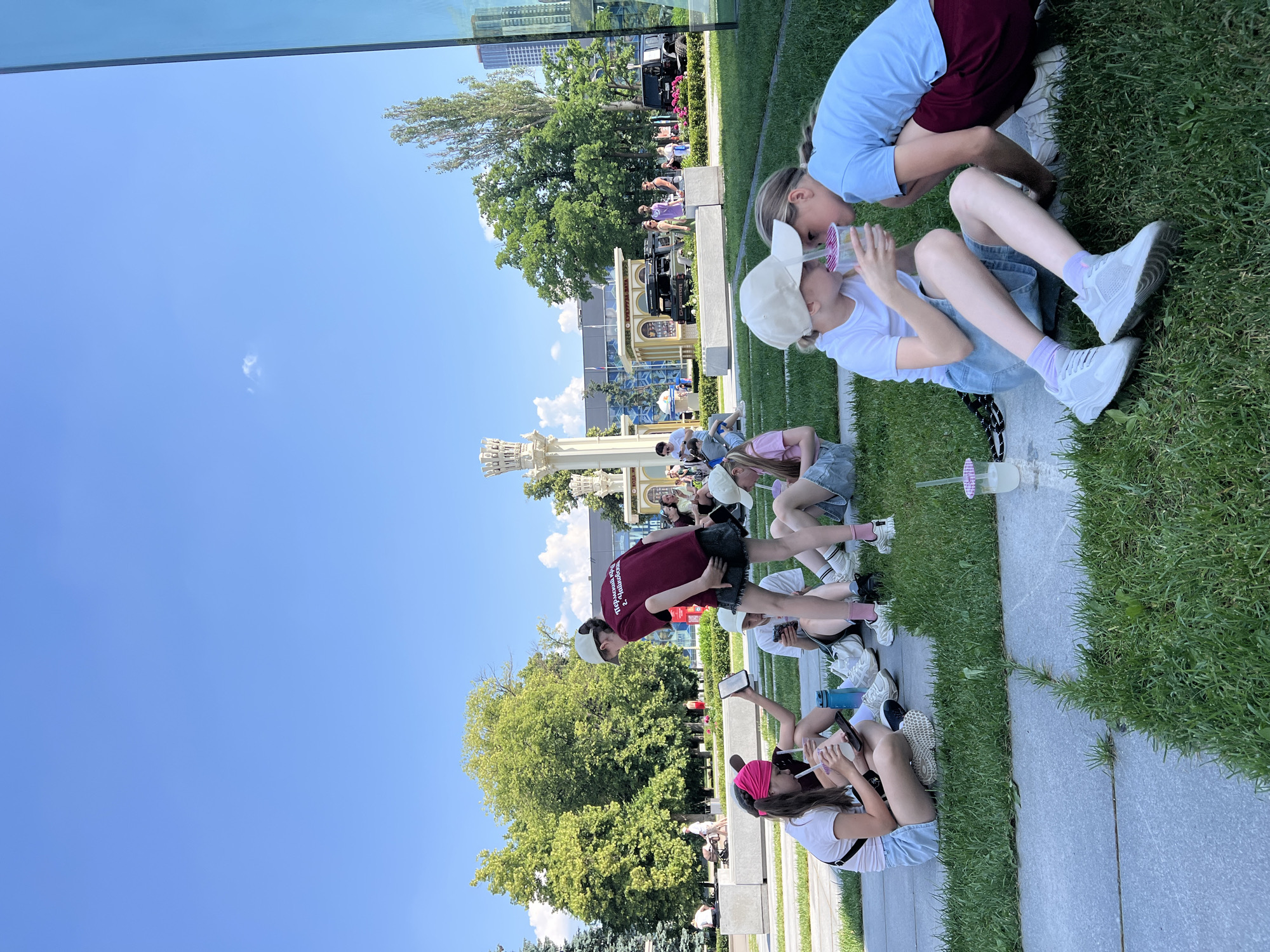 In the foreground, two young girls sit on the grass, one wearing a white t-shirt and denim shorts, the other a light blue t-shirt and maroon shorts. The girl in white is holding a clear plastic cup with a pink lid and straw, and the girl in blue is leaning in, both appearing to be sharing a drink. To their left, another group of girls are also seated on the grass, some with drinks and phones. One girl in a maroon t-shirt and dark shorts stands with her phone in hand. The scene is set in a park-like area with manicured green grass and a stone pathway. In the background, a modern building with a glass facade is visible, as well as trees and a decorative white structure with intricate detailing. Several other people and vehicles are present in the midground and background, suggesting a public outdoor space. The sky is clear and blue. This scene is located in Moscow, Russia, specifically at the VDNKh exhibition center.