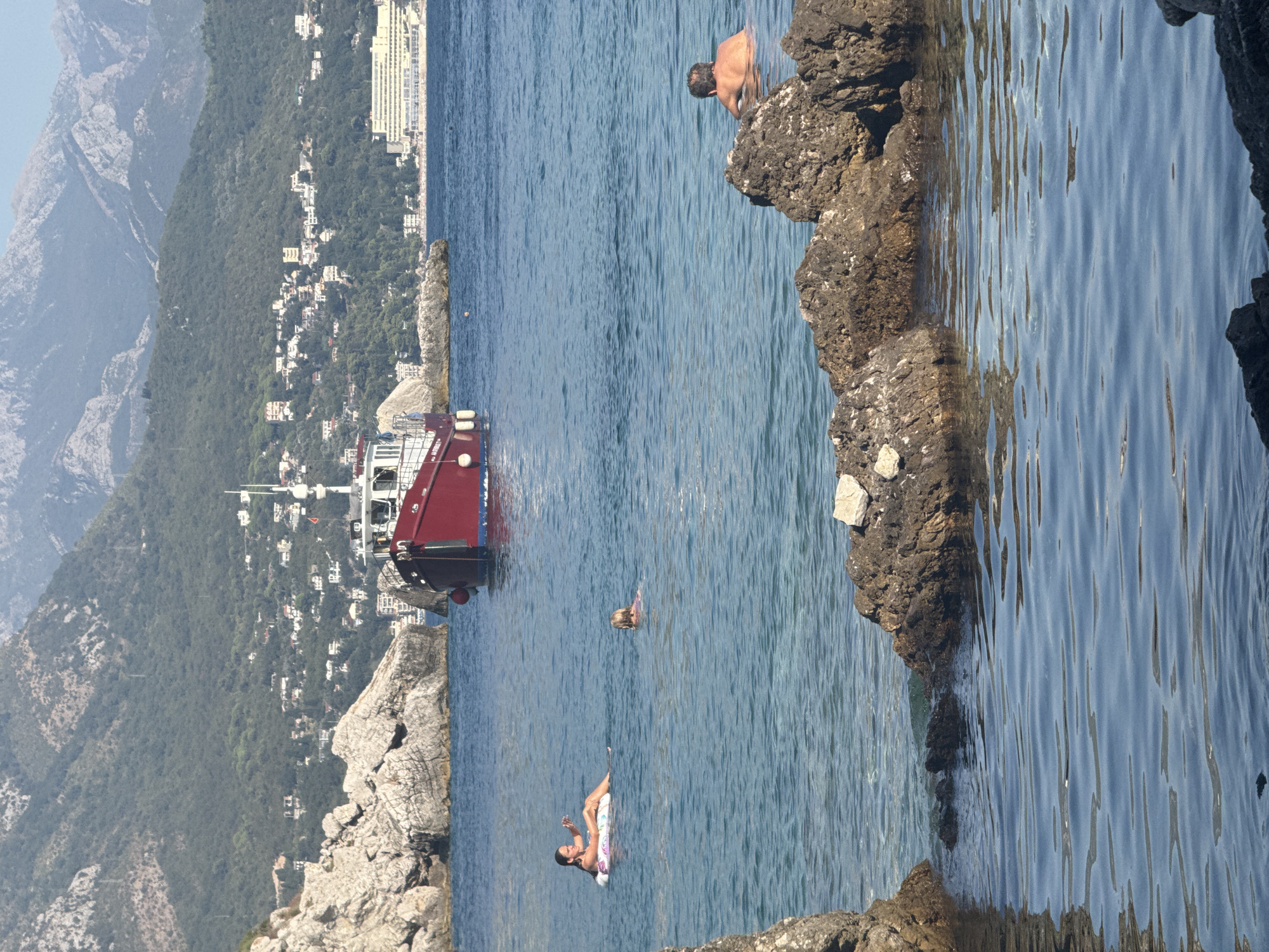 A medium shot captures a serene scene in the waters near Sutomore, Montenegro. In the foreground, dark, rugged rocks break the surface, reflecting the clear blue water. Further out, a red and white boat is anchored. Three individuals are enjoying the water: a woman floats on an inflatable ring, holding a phone; a child swims nearby; and a man, with his back to the camera, wades in the sea. The background features a lush green mountainside dotted with white buildings, suggesting a coastal town. The sea is calm, with gentle ripples on its surface, and the sky is a pale blue.