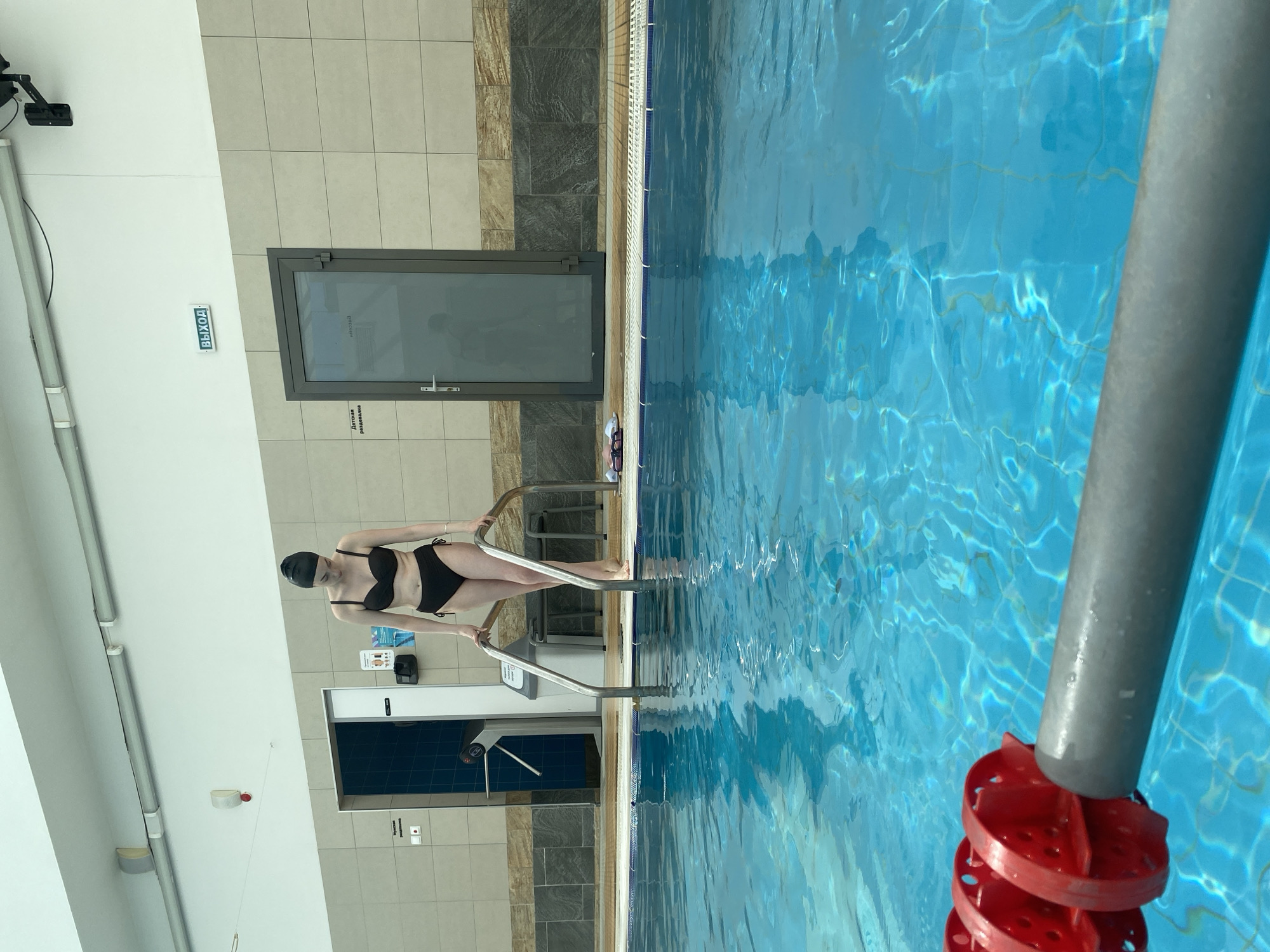 A woman in a black swimsuit and swim cap is standing on a pool ladder, partially submerged in a swimming pool. The pool is indoors, with tiled walls and a door leading to a changing room visible in the background. A sign in Russian, "Выход" (Exit), is visible above the door. A red and gray pool divider is in the water on the right side of the frame. The photo appears to be taken from a slightly elevated position. The location is likely a swimming pool facility in Yekaterinburg, Russia.