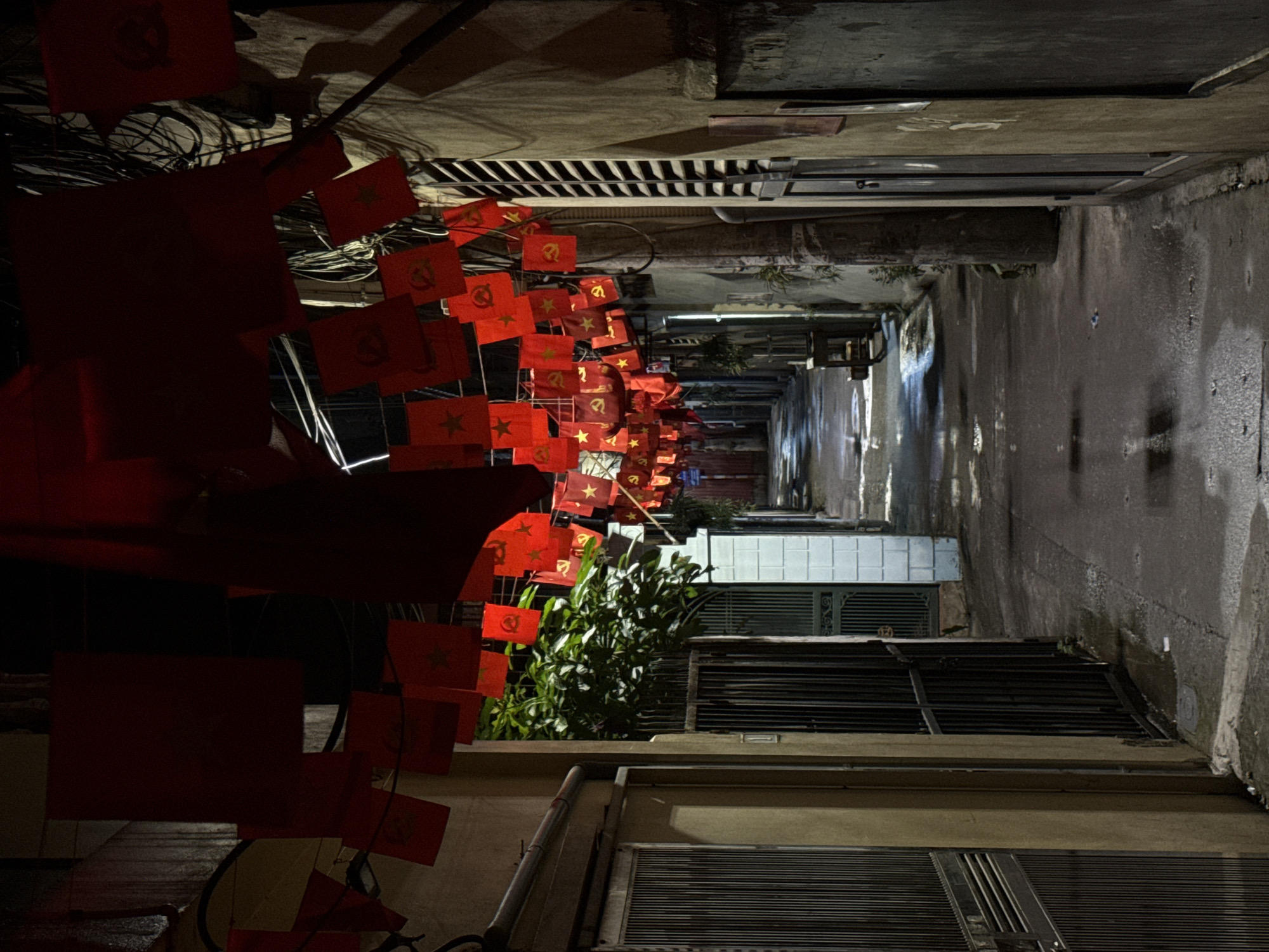 The image depicts a narrow alleyway in Hà Nội, Vietnam, decorated with numerous small red flags bearing the Communist hammer and sickle emblem, or a star. These flags are strung across the alley's entrance, creating a dense, visually striking effect. The alley itself is dimly lit, with wet pavement reflecting the sparse light. Buildings flank both sides of the alley, showing closed window shutters and aged walls. Electrical wires are tangled above the flags, adding to the cluttered urban scene. The perspective is from within the alley, looking toward the brighter opening at the far end. No people are visible in the image.