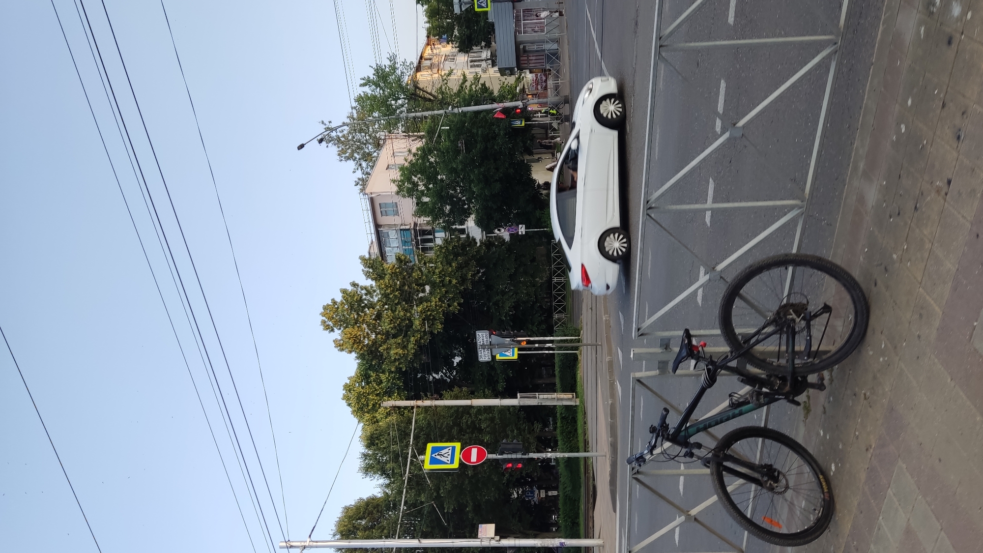 The image depicts a street scene in Krasnodar, Russia, during daylight. In the foreground, a black mountain bike is parked on a sidewalk next to a metal barrier. The bicycle has a green frame and appears to be in good condition. Beyond the barrier, a white sedan is captured in motion, turning at an intersection. Traffic lights and pedestrian crossing signs are visible. The background reveals multi-story buildings with residential apartments and mature trees lining the street. Overhead power lines are strung across the sky. The sky is clear and blue, suggesting fair weather. The overall impression is of a typical urban environment.