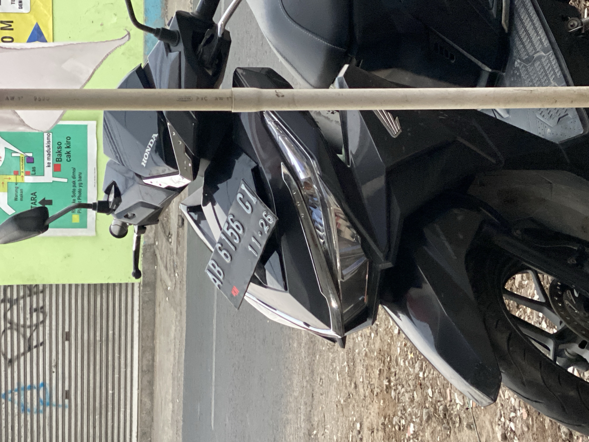 The image shows a black Honda motorcycle parked outdoors on a street in Kasihan, Indonesia. The motorcycle is the primary subject, with its front half prominently displayed. It has a license plate that reads "AB 6156 CI" and a date stamp of "11-26". The motorcycle is parked on a gravelly shoulder next to a paved road. In the background, there is a green building with a metal roll-up door that has blue graffiti on it. A map is also visible on the side of the building. A white pipe, likely part of a temporary structure or utility, runs vertically through the center of the frame, partially obscuring the motorcycle. The lighting suggests it is daytime.