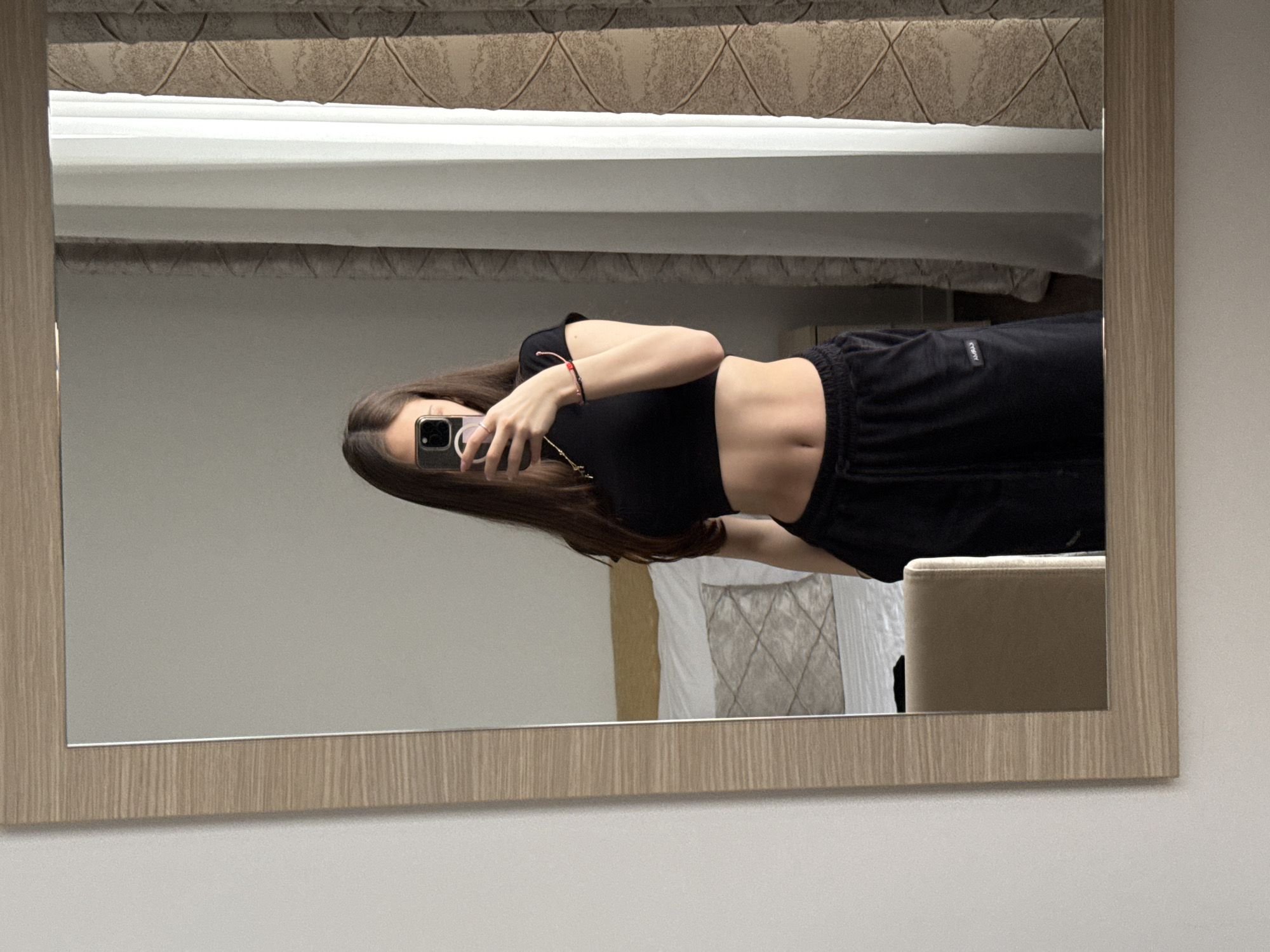 A young adult female is depicted taking a mirror selfie within a room in the Vozdovac Urban Municipality, Serbia. She has long, dark brown hair and is wearing a black crop top exposing her midriff, paired with black sweatpants featuring a drawstring and a small white "AMBAS" label on the left leg. Her right hand holds a black smartphone with a clear case, partially obscuring her face; a silver-colored ring is visible on her right ring finger. She wears a thin gold-colored chain necklace with a small pendant and multiple bracelets on her left wrist, including one red string bracelet. The mirror, framed in light-colored wood with a horizontal grain, reflects a bedroom environment behind her. This includes a bed with a light-colored wooden headboard, white linens, and a patterned grey pillow. To the right, a window with sheer white and patterned beige curtains is visible. A portion of a light-colored, possibly beige, upholstered chair is reflected in the lower left segment of the mirror. The mirror is mounted on a plain white wall.