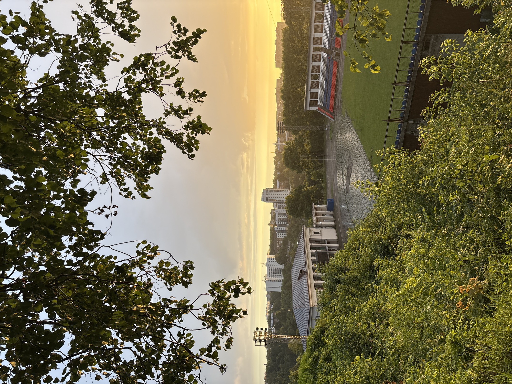 This is a high-angle, wide shot of a stadium in Sof'ino, Russia, during sunset. The sky is a vibrant orange and yellow, with some clouds. In the foreground, lush green trees and bushes partially obscure the view. Below the trees, a building with a gray, corrugated roof and white walls is visible. Adjacent to this building is a football field with green grass and a wet, paved area. Stadium seating, in red, white, and blue, lines one side of the field. In the background, a cityscape with white, multi-story buildings and a large light tower can be seen. The overall atmosphere is serene and picturesque.