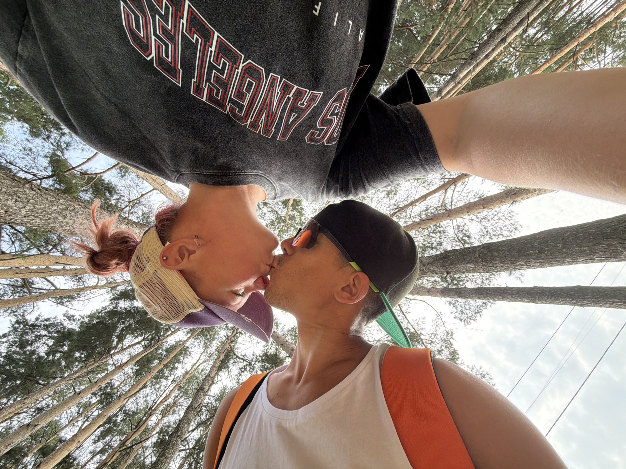 Two individuals are captured in a close-up, low-angle selfie, appearing to share a kiss amidst a backdrop of tall trees. The image is rotated, with the subjects positioned at the top.
The person on the left, whose head is tilted back, has pink-hued hair styled in a bun and is wearing a dark grey t-shirt with red and white lettering that reads "LOS ANGELES". They are also wearing a mesh trucker hat with a purple brim. Visible on their ear is an earring.
The person on the right is wearing a white tank top with an orange backpack. They have a dark baseball cap with a neon green brim and are wearing sunglasses. Their left arm is visible, and they have facial hair.
The environment consists of numerous tall pine trees with green foliage, suggesting an outdoor setting, possibly a forest or park. The sky is visible through the branches, with some clouds present. The image gives the impression of being taken during daylight hours. The context of Chaikovsky, Russia, is not visually ascertainable from the image itself.