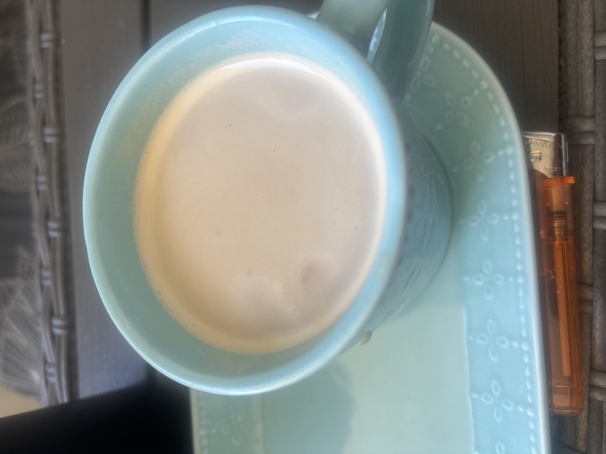 An overhead close-up shot shows a light blue ceramic mug filled with a beige liquid, likely coffee with milk. The mug sits on a matching light blue saucer with a decorative embossed pattern of small, four-petal flowers and dots. In the foreground, a small orange disposable lighter is partially visible, resting on the saucer. The background is blurred but appears to be an outdoor setting with a woven textured surface. The scene is not identifiable as being in Plovdiv, Bulgaria.