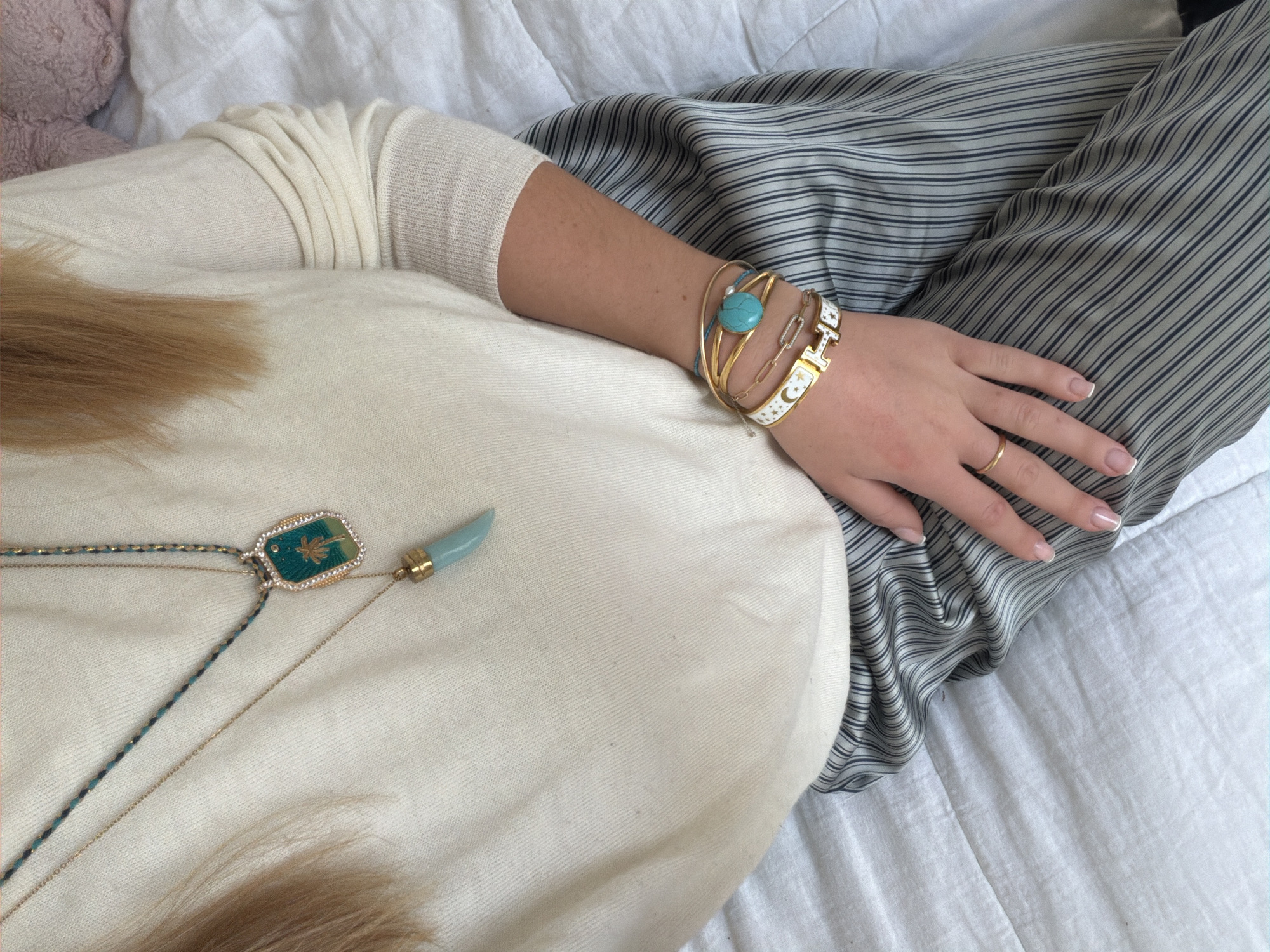 An indoor scene, located in Paris, France, depicts a person from an overhead perspective, partially visible from the chest to the mid-thighs. The individual has fair skin and light-colored hair, visible on the left side of the frame. They are wearing a cream-colored, long-sleeved sweater and grey pants with thin black vertical pinstripes.
Two necklaces are visible: a shorter gold chain featuring a light blue horn-shaped pendant, and a longer braided green and gold cord necklace suspending a rectangular pendant with a gold frame, white rhinestones, and a central golden palm tree motif against a teal background.
The person's right arm and hand are prominent, with the hand resting on their leg. The wrist is adorned with a stack of bracelets, including multiple thin gold bangles, a gold paperclip chain bracelet, a bracelet with a large, round turquoise stone, and a white enamel bracelet detailed with gold stars and a central 'H' logo. A plain gold ring is present on the right ring finger, and the fingernails exhibit a French manicure style. The individual is situated on a white, quilted soft surface, likely a bedspread. A partial view of a soft, pink object is visible in the upper right background.