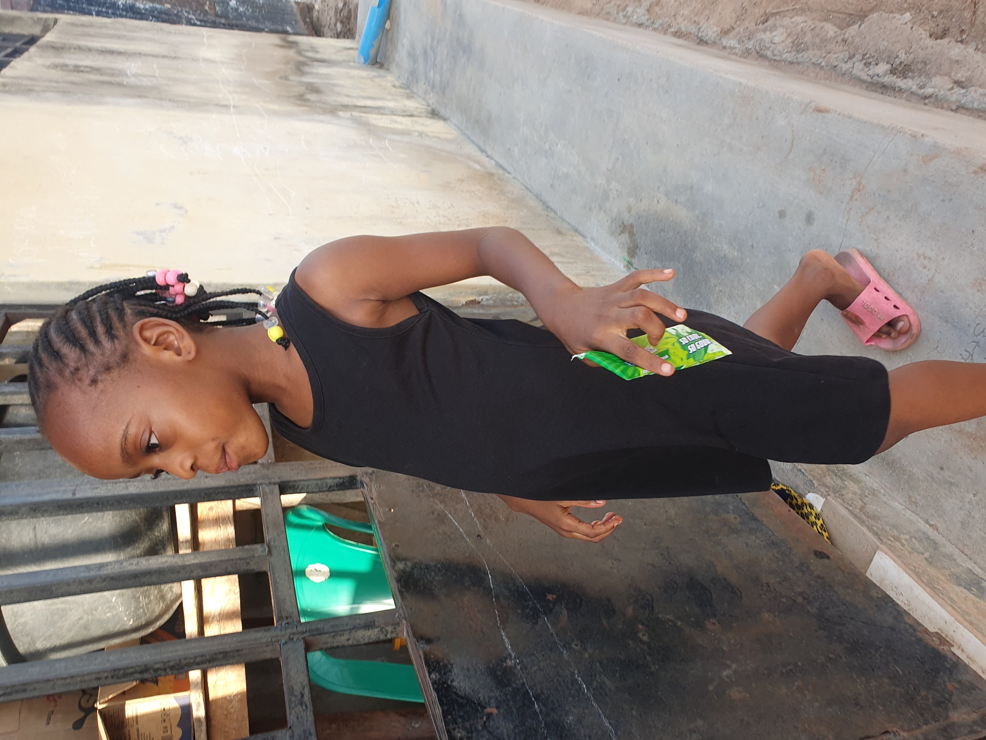 A young girl is standing on a concrete surface in an outdoor environment. She is wearing a black dress and pink sandals. Her hair is braided with colorful beads. She is holding a green and white packet. A metal structure and various objects, including a green plastic item, are partially visible nearby. The location is suggested to be in the city of Lokoja, Nigeria.