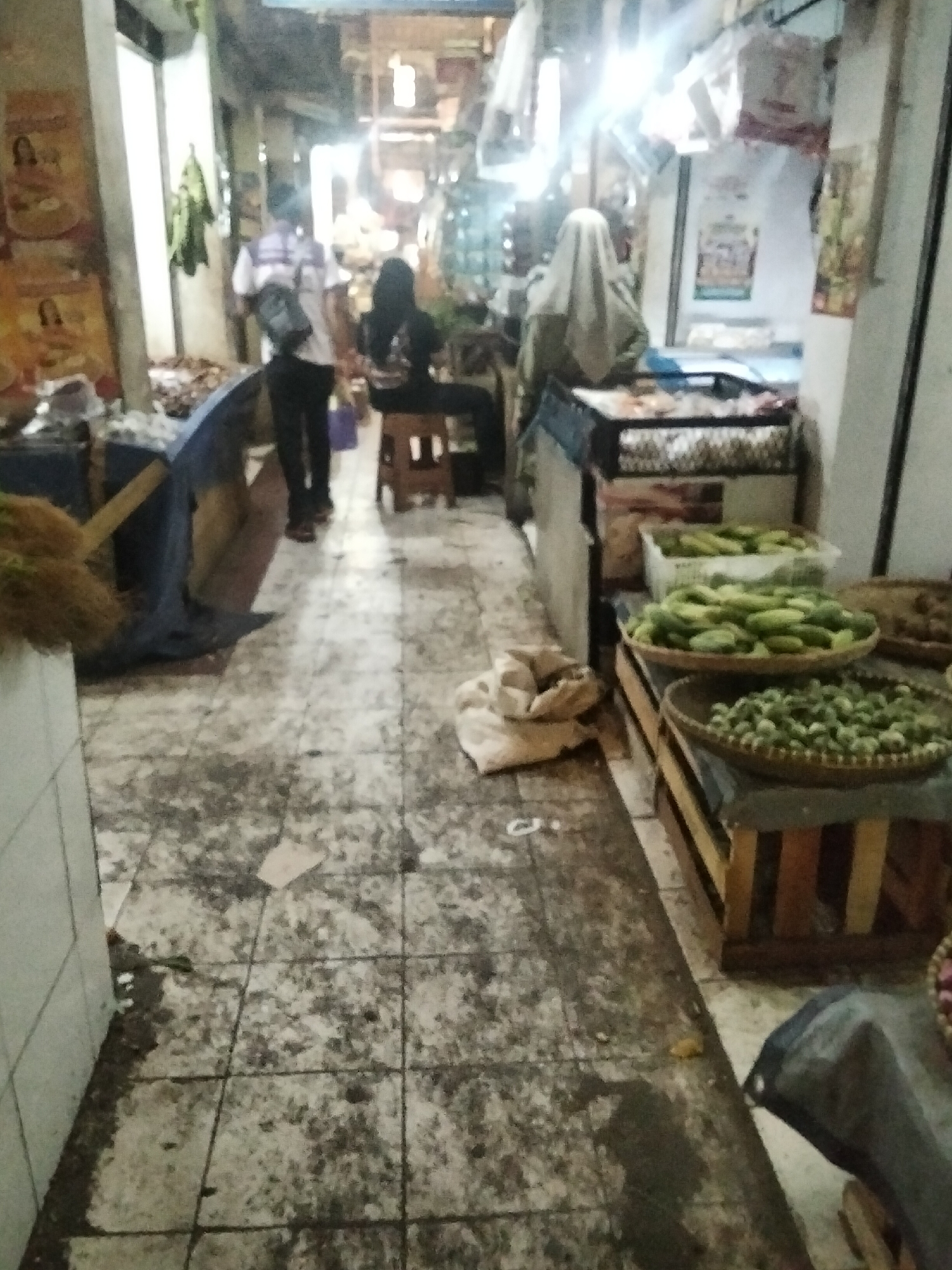The image depicts a busy market scene in Cisaat, Indonesia. The floor is tiled and appears dirty and worn, with debris scattered across it. To the left, a person in a white and striped shirt, dark pants, and a dark bag slung across their shoulder, walks away from the camera. Beside them, a dark blue tarp is draped over an unseen object, with straw-like material protruding from its left side. Further down the path, a person sits on a wooden stool wearing a dark headscarf and dark clothing. On the right side of the image, various produce is displayed. A large woven basket holds numerous green cucumbers. Below it, another smaller basket contains what appear to be small green vegetables. Behind these baskets, a wooden crate holds more green produce. In the background, several people are visible, including one person wearing a white head covering, standing behind a counter. The market appears to be indoors with artificial lighting illuminating the space, creating bright spots and shadows. Posters are visible on the walls.