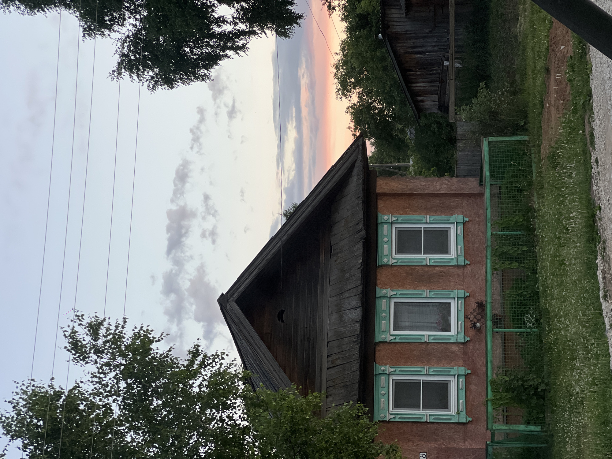 This is a photograph of a weathered, wooden house with red-brown stucco walls, located in the village of Pigozino, Russia. Three windows with ornate, teal-colored frames and shutters are visible on the front facade. A low, green metal fence encloses a small yard with overgrown grass and wildflowers. The roof of the house is made of dark, aged wood. In the background, a darker wooden structure, possibly a shed or another house, is visible, along with lush green trees and a hint of a pastel-colored sunset in the sky. Overhead, power lines stretch across the frame. No people or animals are present in the image.