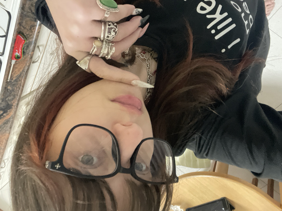 An overhead-angle self-portrait depicts an individual indoors, identified as being in Petah Tikva, Israel. The person has long, dark brown hair with reddish highlights and bangs, wearing black-framed glasses with clear lenses and a small cross emblem on the upper left frame. Their light-colored eyes are directed towards the camera. They have light pink lip color. The individual wears a black long-sleeved shirt with white text on the chest, partially visible as "i like". Their left hand (viewer's right) is raised, with the index finger touching their chin. This hand displays multiple rings: a large plain ring on the thumb, two rings on the index finger (one with an oval stone), two ornate rings on the middle finger, and a ring with a large green oval stone on the ring finger. The fingernails are long and pointed; those on the left hand are white with glitter, while those on the right hand are dark with white elements. A silver-colored chain necklace is visible around their neck.
The background shows an indoor setting. To the upper right, a dark, speckled kitchen countertop is visible, with a silver gas stovetop burner and a red and white lighter resting on it. To the lower left, a light-colored wooden table, possibly round, and a wooden chair are present. A dark mobile phone lies on the wooden table. The floor appears to be light-colored tiles.