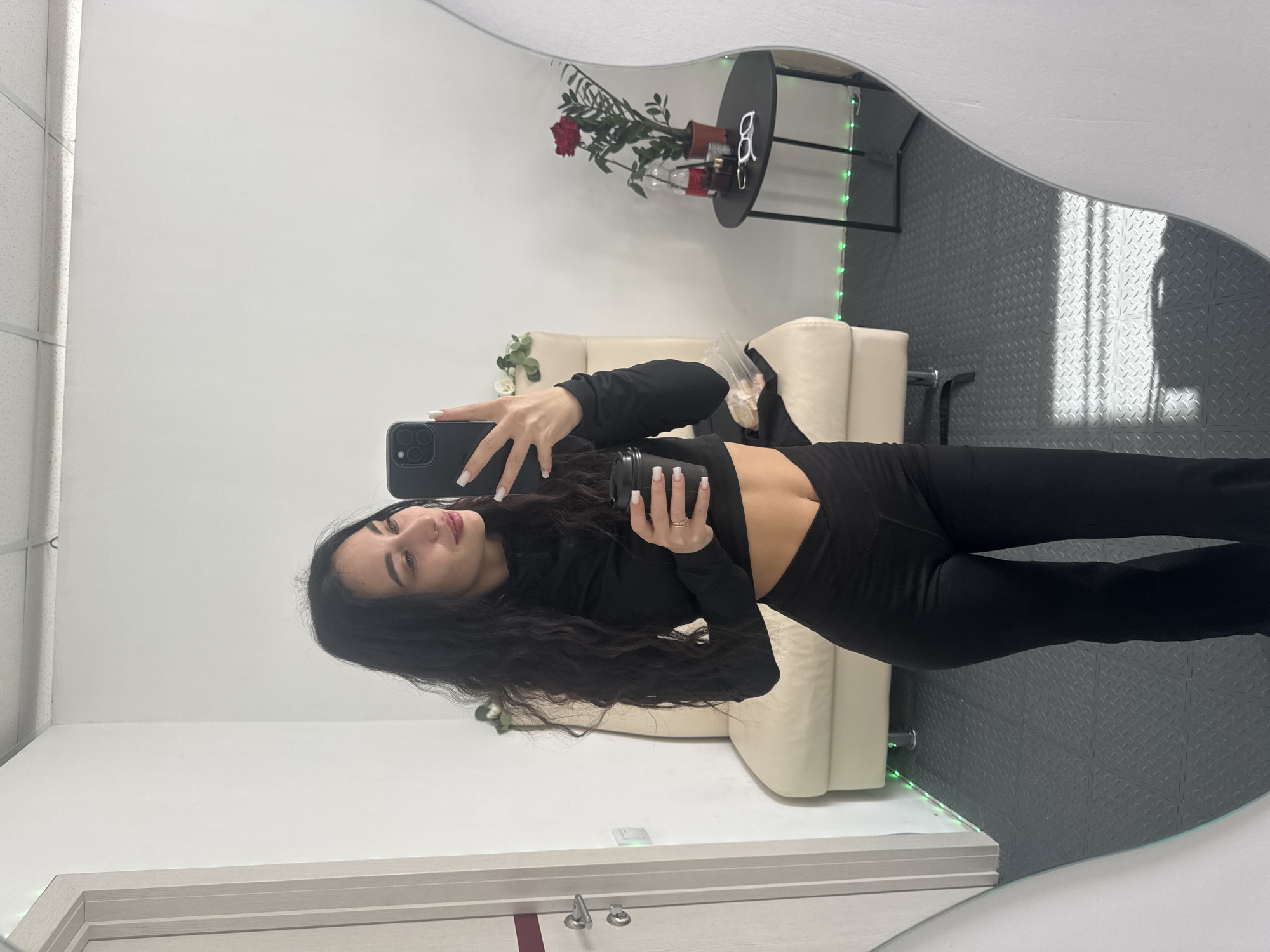 A young woman is taking a selfie in a mirror. She is wearing a black long-sleeved crop top and black leggings. She is holding a black cell phone in one hand and a black cup or container in the other. The mirror is reflecting a room with a white wall, a beige couch, a black table with a plant and some other items on it, and a gray patterned floor. Based on the provided information, the location of the room is in the city of Фоки, Russia.