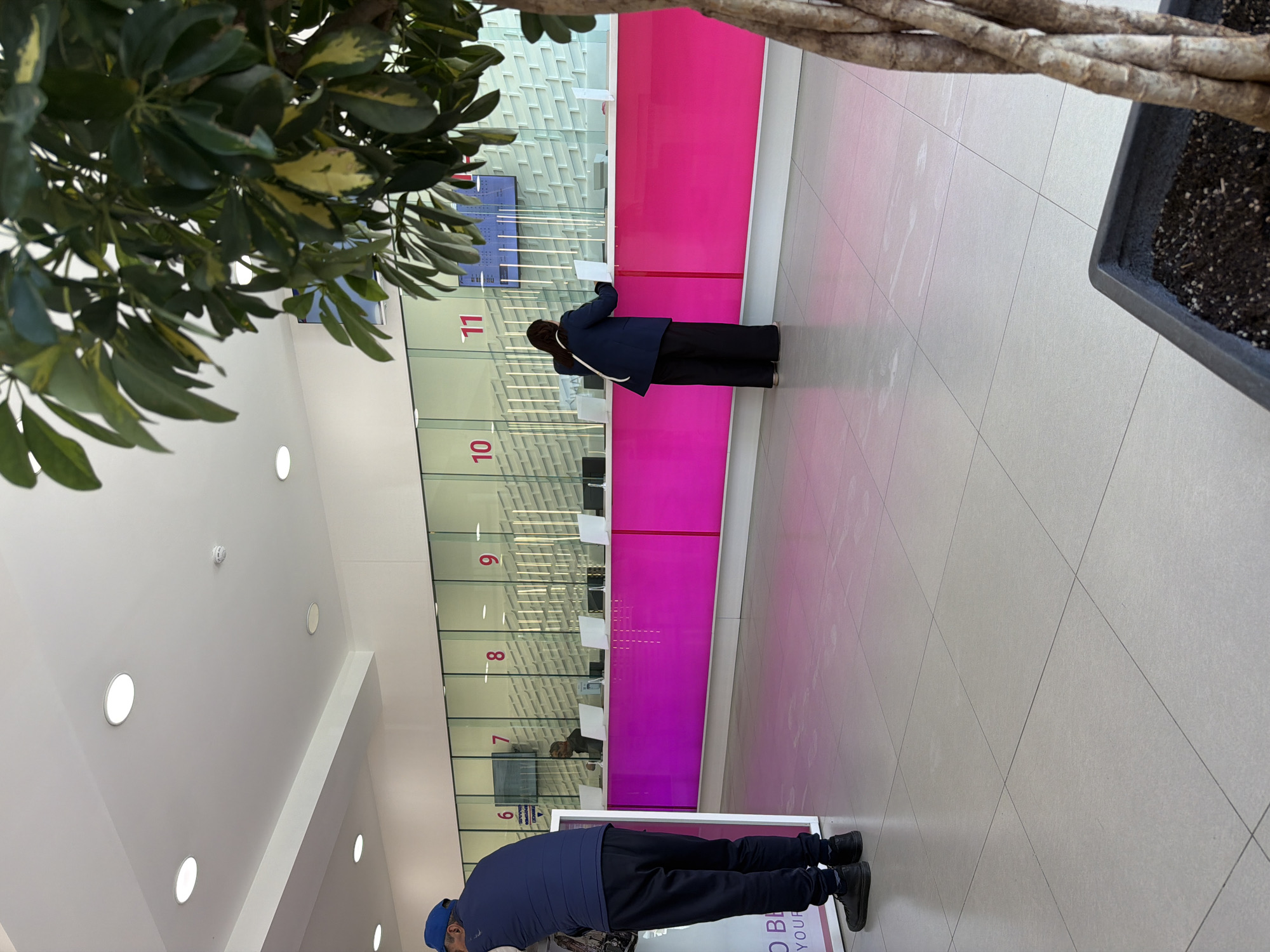 The image shows an indoor scene, likely a public service area, in what could be a branch of HayPost in Yerevan, Armenia. There is a service counter with numbered stations, marked from at least 6 to 11. The counter is painted in a vivid pink and white with glass partitions. Two individuals are seen at the counter. One person, wearing a dark blue jacket and black pants, is standing at station 11, facing the counter, appearing to be filling out a document or interacting with the staff behind the window. Another person, wearing a blue top, black pants and a blue cap, is standing at station 6. The floor is tiled with light grey tiles with a diamond pattern. A tree is partially visible on either side of the frame. The ceiling is white with several recessed circular lights. The setting suggests a modern, renovated interior.