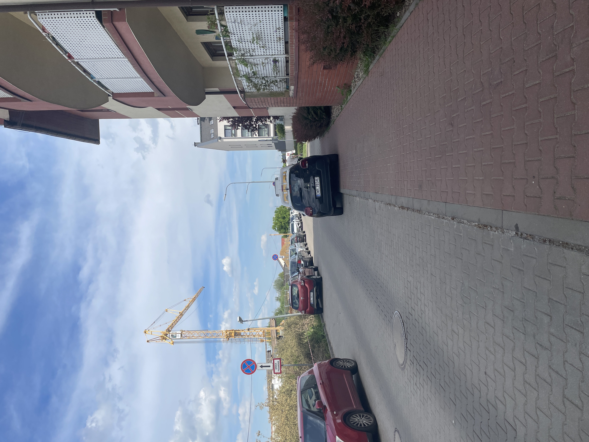 The image shows a street in Wysoka, Poland. Several cars are parked along the side of the road, including a black Citroen with license plate DMR 7834U and a red Suzuki. A construction crane is visible in the background, under a blue sky. A street sign is visible. The road is paved with gray and brown pavers. The buildings are modern with balconies.