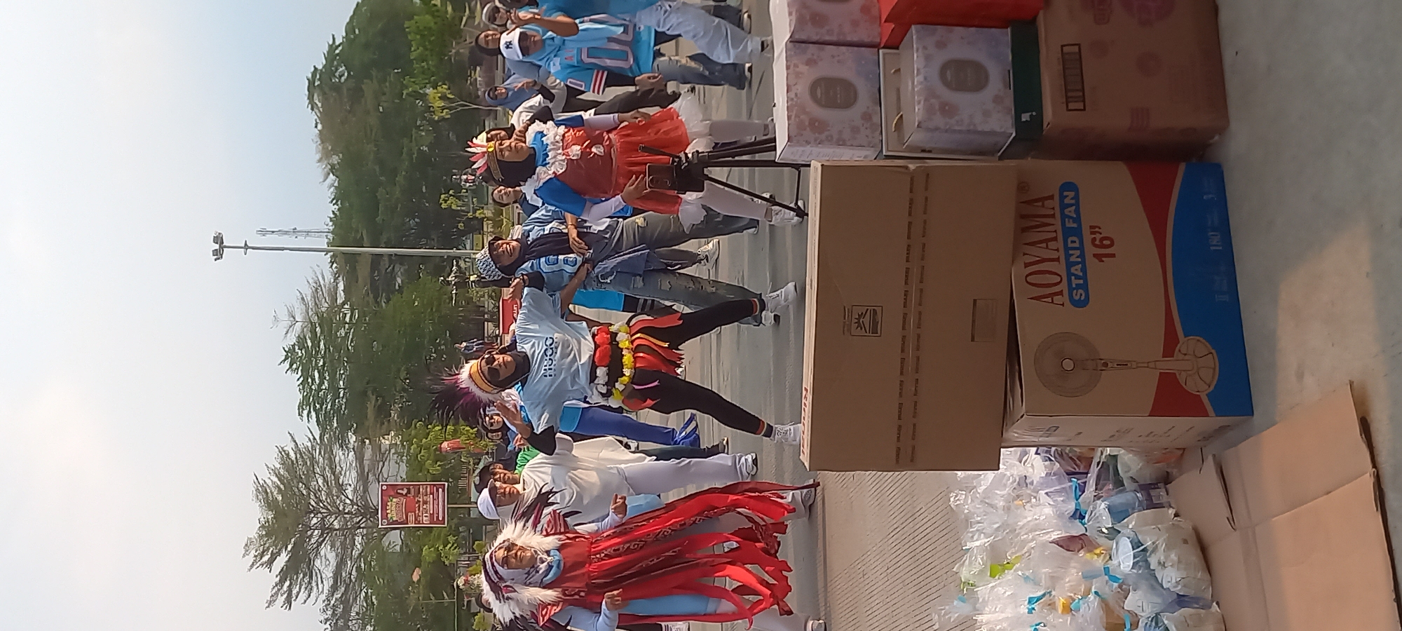 In Melayu, Indonesia, a group of women is engaged in an outdoor activity, possibly a dance or exercise session. They are dressed in various costumes, with some wearing elaborate headwear resembling Native American headdresses. One woman in the foreground wears a light blue t-shirt with "HUGO" printed on it, along with a colorful fringed skirt. Another woman to her left is adorned in a red and blue costume with dangling fabric strips, also wearing a headdress. The background features trees, a lamppost, and a billboard. In the lower portion of the image, several cardboard boxes are stacked, including one with the text "AOYAMA STAND FAN 16"" visible. Plastic-wrapped items are also scattered on the ground. The overall scene suggests a public event or gathering taking place in an urban or semi-urban outdoor space.