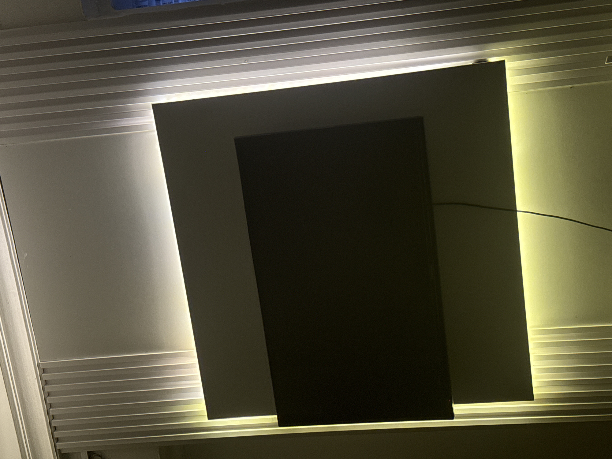 A modern living room in Abuja, Nigeria, features a large, dark-colored television mounted on a textured white wall. The television is recessed into a slightly larger rectangular panel, which is offset from the main wall. Integrated LED lighting illuminates the space from behind the rectangular panel, casting a warm yellow glow. The surrounding wall is decorated with vertical fluted panels, adding a subtle architectural detail. The lighting highlights the textures of both the television panel and the fluted wall.