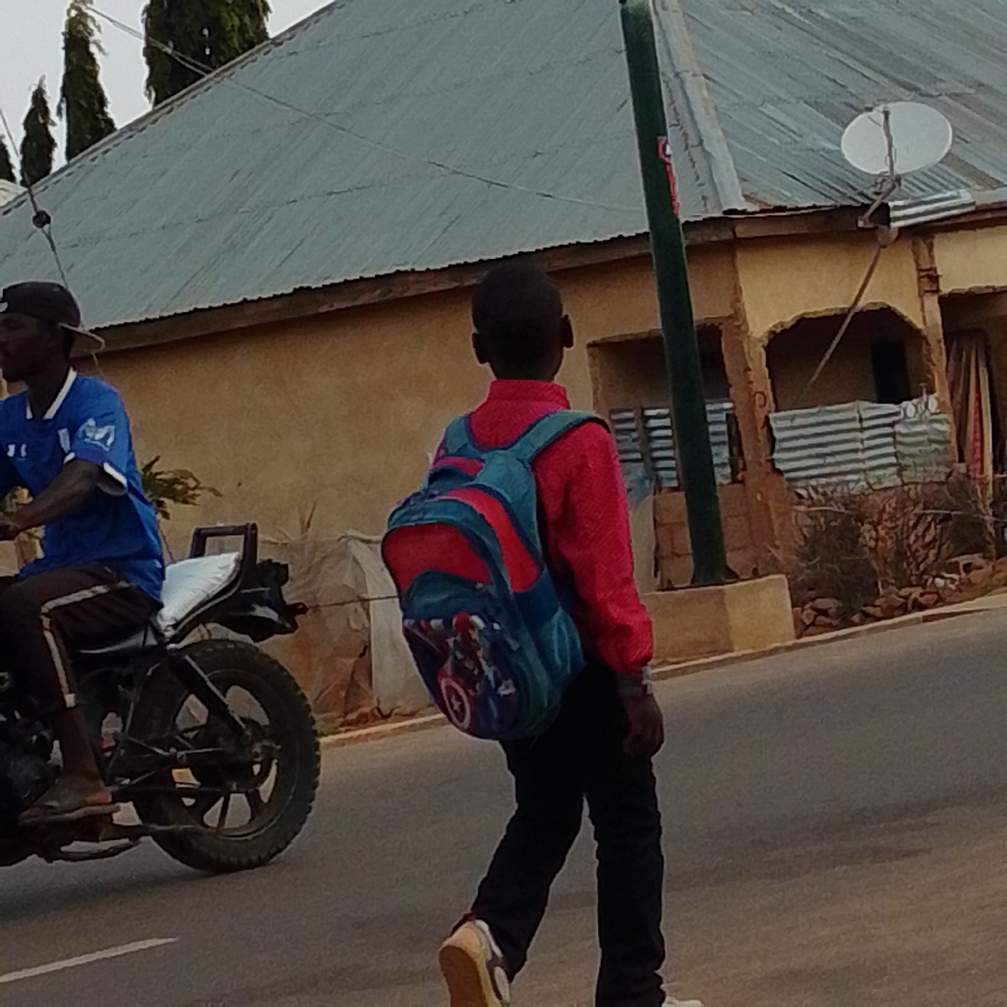 In Gombe, Nigeria, on a paved road, a young boy walks with a blue and red backpack adorned with superhero imagery. He wears a bright red long-sleeved shirt and black pants. To his left, a man rides a motorcycle. He is wearing a blue t-shirt and a dark baseball cap. The background features a building with a corrugated metal roof and a satellite dish. The sky is overcast.