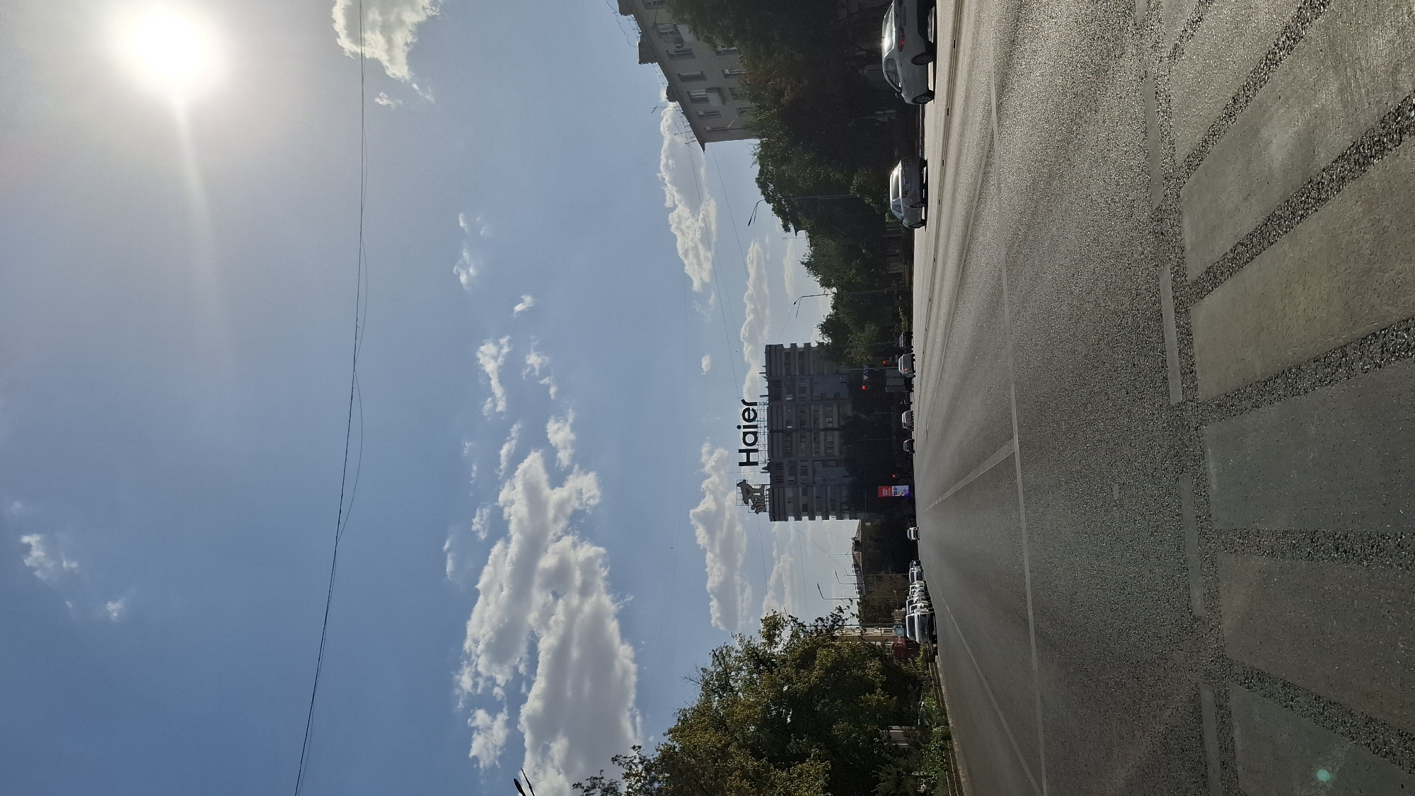 The image depicts a street scene in Shymkent, Kazakhstan, likely on a sunny day with scattered clouds. The foreground features an asphalt road with clear lane markings and a pedestrian crossing. On the left side of the road, parked cars are visible along the sidewalk, partially obscured by trees.
In the mid-ground, several cars are driving on the road. Dominating the background is a multi-story building with a prominent "Haier" logo on its roof. Adjacent to this building, on the right side of the image, is another building with a similar architectural style. Trees line the sides of the road, contributing to the urban landscape. The bright sun is positioned in the upper right corner of the frame, casting a glare.