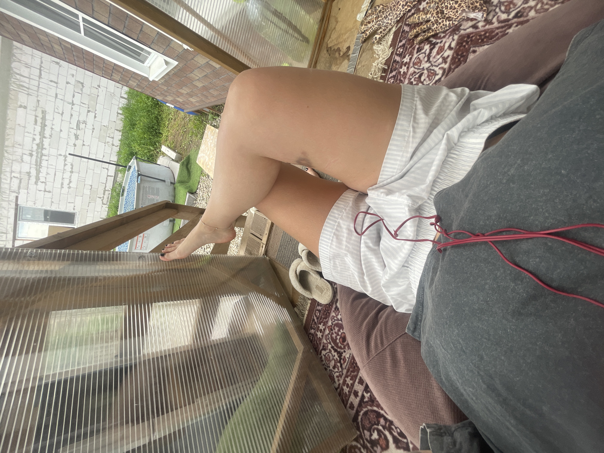 The image shows a person indoors, likely resting. The lower half of the person is visible, revealing bare legs crossed at the knees and covered with a white, striped garment. The person is lying on a cushion. Red cords are draped across the clothing. The environment is cluttered, with a structure made of wood and translucent sheeting, suggesting a porch or sunroom. Outside the structure, a house with visible white brickwork and a rectangular window is seen. Green vegetation and a small pool or tub are also visible in the background. The location is near Мосметаллоконструкция Онт, Russia.