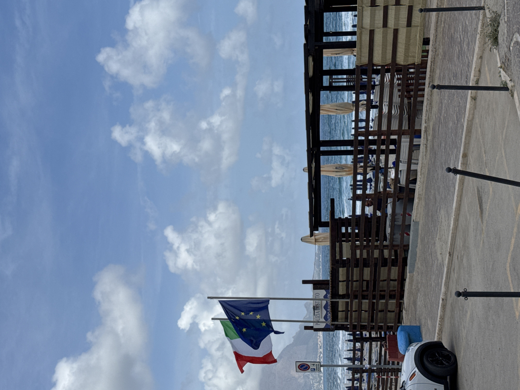 The image depicts a view of a beach establishment on the coast of Isola delle Femmine, Italy. In the foreground, a paved area with parking bollards is visible, with a white car parked on the left. Beyond the paved area, a wooden fence encloses a beach facility with rows of folded chairs and umbrellas. Two flags, the Italian flag and the European Union flag, are prominently displayed on flagpoles to the left of the beach establishment. In the background, the sea is visible with moderate waves, and a mountainous landscape can be seen in the distance under a cloudy sky. A sign near the flags reads "POLIZIA DI STATO" and "CENTRO BALNEARE".