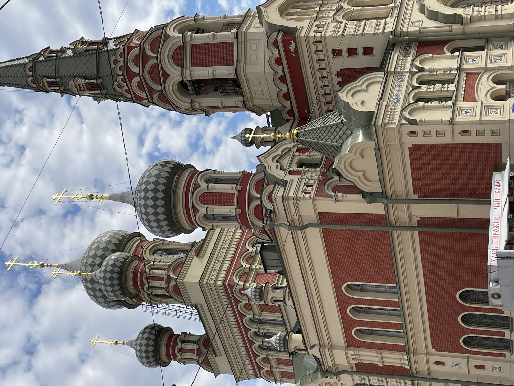 The image captures a grand, ornate Russian Orthodox church under a bright, partly cloudy sky. The primary subject is the multi-domed and spired church, characterized by its distinctive red brickwork contrasting with cream-colored stucco and intricate decorative elements. Several onion-shaped domes, featuring a gray and white checkered pattern, are visible, each topped with a golden, multi-barred Orthodox cross. A tall, slender spire also rises prominently, adorned with similar architectural details and capped with a golden cross.
The building's facade showcases a rich array of architectural features, including arched windows, decorative kokoshniks, and tiled patterns integrated into the cream-colored trim. The lighting suggests it is daytime. Overhead, thin electrical wires or cables traverse the scene.
On the lower right side of the image, a partially visible white sign displays Russian text. The upper line reads "ХЛЕБ ПИРОЖКИ ЧАЙ К" (BREAD PIES TEA K), and a lower line clearly shows "КОФЕ" (COFFEE), indicating a nearby establishment such as a bakery or cafe. The intricate and colorful architecture is consistent with historical buildings found in Saint Petersburg, Russia.