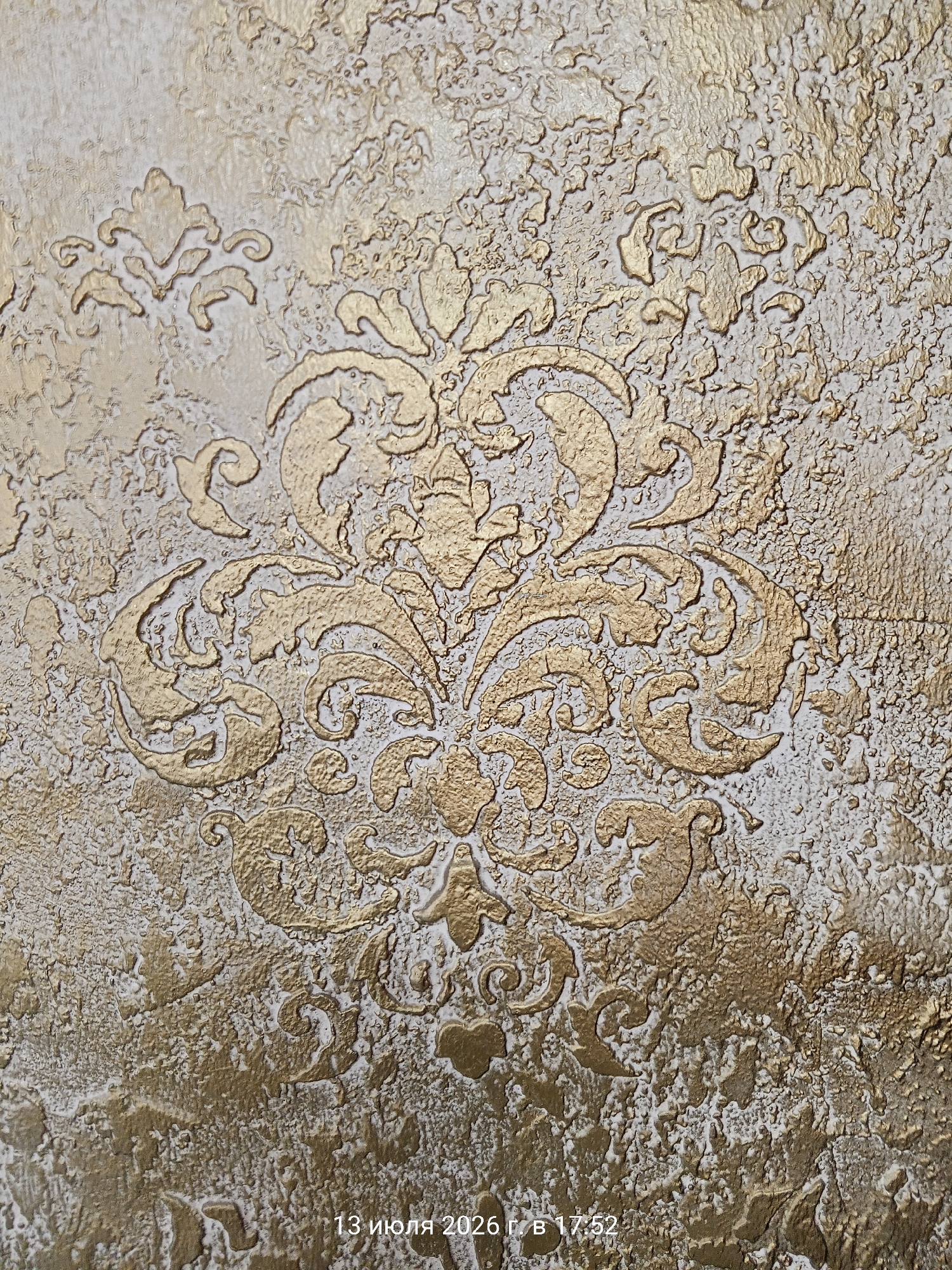 The image shows a close-up of textured wallpaper with a damask-like pattern in gold and beige. The wallpaper has a stucco-like finish with raised floral and swirling motifs highlighted in gold. The texture is rough and uneven, giving a three-dimensional effect. In the bottom right corner, there is text that reads "13 июля 2026 г. в 17:52". There are no people, actions, or interactions visible in the image. The location is specified as Ust-Dzheguta, Russia, but this information is not visually evident from the photograph itself.