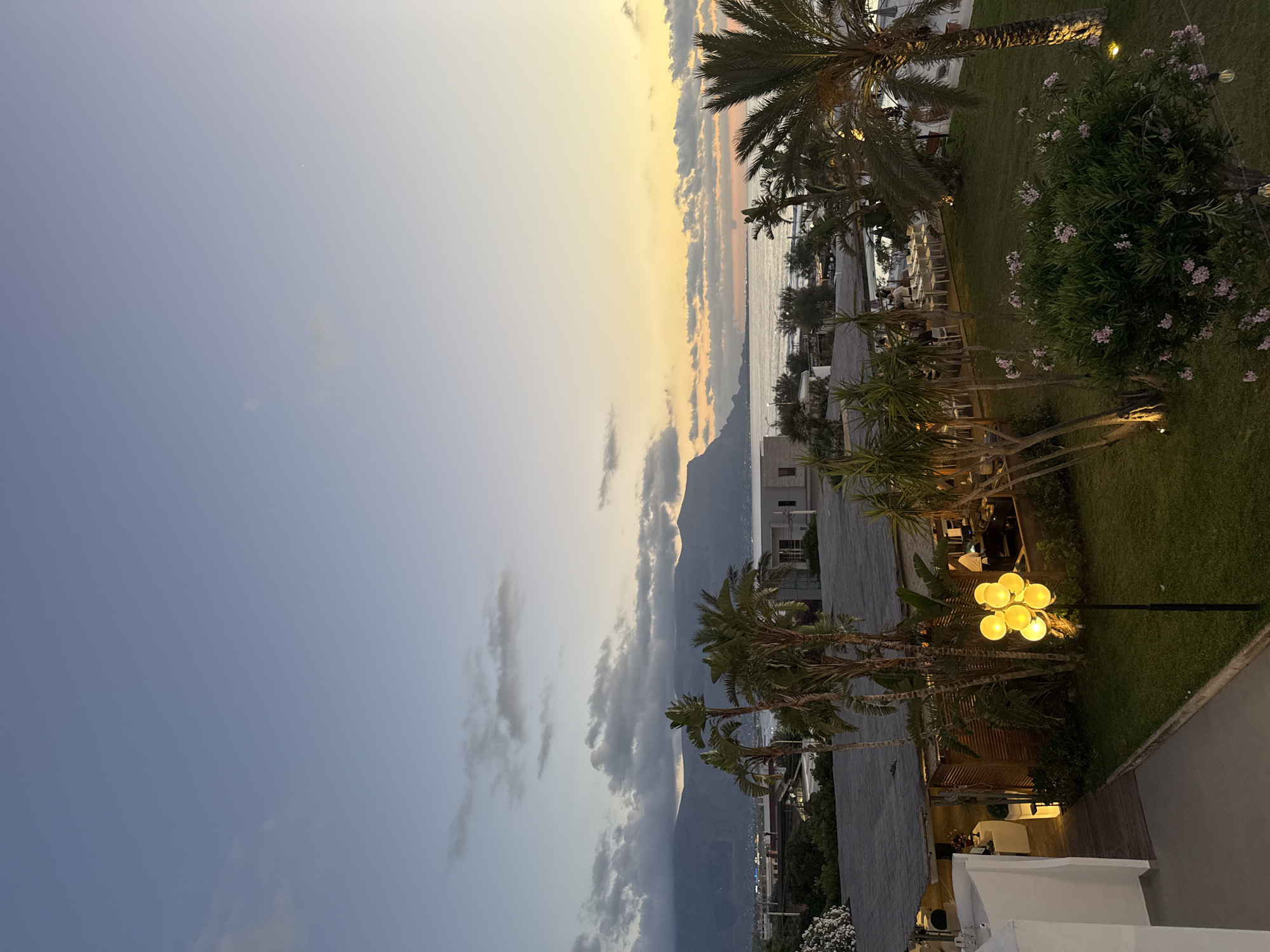 This is a medium-shot, eye-level, outdoor photograph taken at dusk overlooking a coastal area in Isola delle Femmine, Italy. The sky transitions from a pale blue at the top to soft yellow and orange hues near the horizon, with scattered clouds. In the distance, a range of mountains frames the coastline, with a town or village visible along the shore. The sea is visible, meeting the land.
In the foreground and midground, there is a restaurant area with outdoor seating, visible beneath a covered structure with a thatched roof. White tables and chairs are arranged on the patio. Palm trees and other lush green foliage are prominent throughout the scene. A cluster of yellow globe lights on a lamp post illuminates a grassy area in front of the restaurant. A low-lying white building is visible on the left side of the frame, with a wooden walkway leading towards the restaurant. In the background, near the sea, there are more trees and some buildings. The lighting suggests it is either sunrise or sunset, with artificial lights from the restaurant and street lamps casting a warm glow. There are no distinct people clearly visible or engaged in discernible actions.