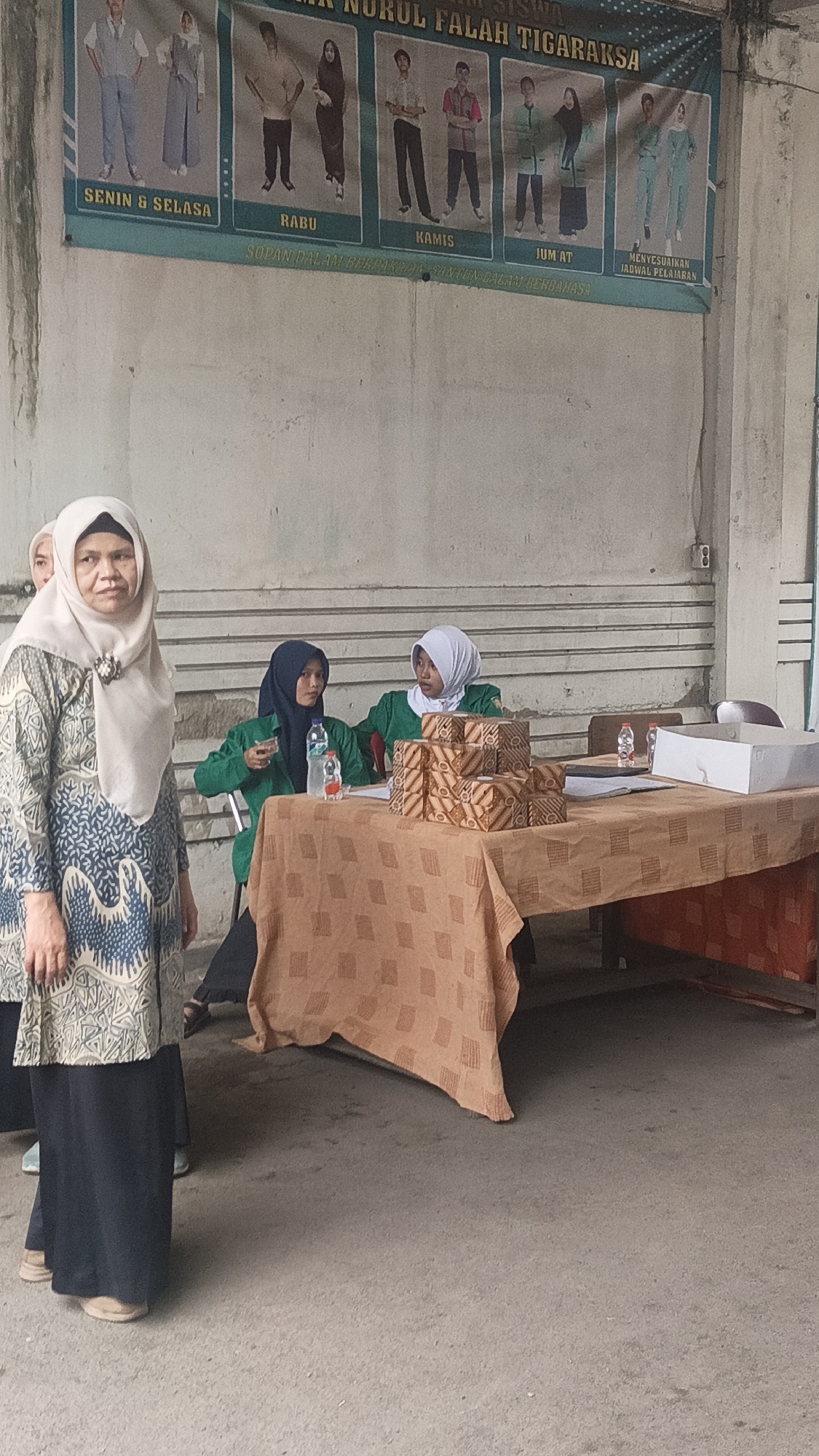 The image captures an outdoor scene in Tigaraksa, Indonesia, likely on school grounds given the banner displayed. In the foreground, a woman wearing a light-colored hijab and a patterned dress stands looking towards the camera. Behind her, two young women wearing green jackets and hijabs are seated at a table covered with an orange patterned tablecloth. One of the seated women is holding a plastic bottle. On the table, there are stacks of boxes with a batik pattern and a white box. The background features a concrete wall with a large banner depicting different uniform styles for students on various days of the week, with text in Indonesian. The ground is paved with concrete. The overall atmosphere appears to be a casual gathering or event.