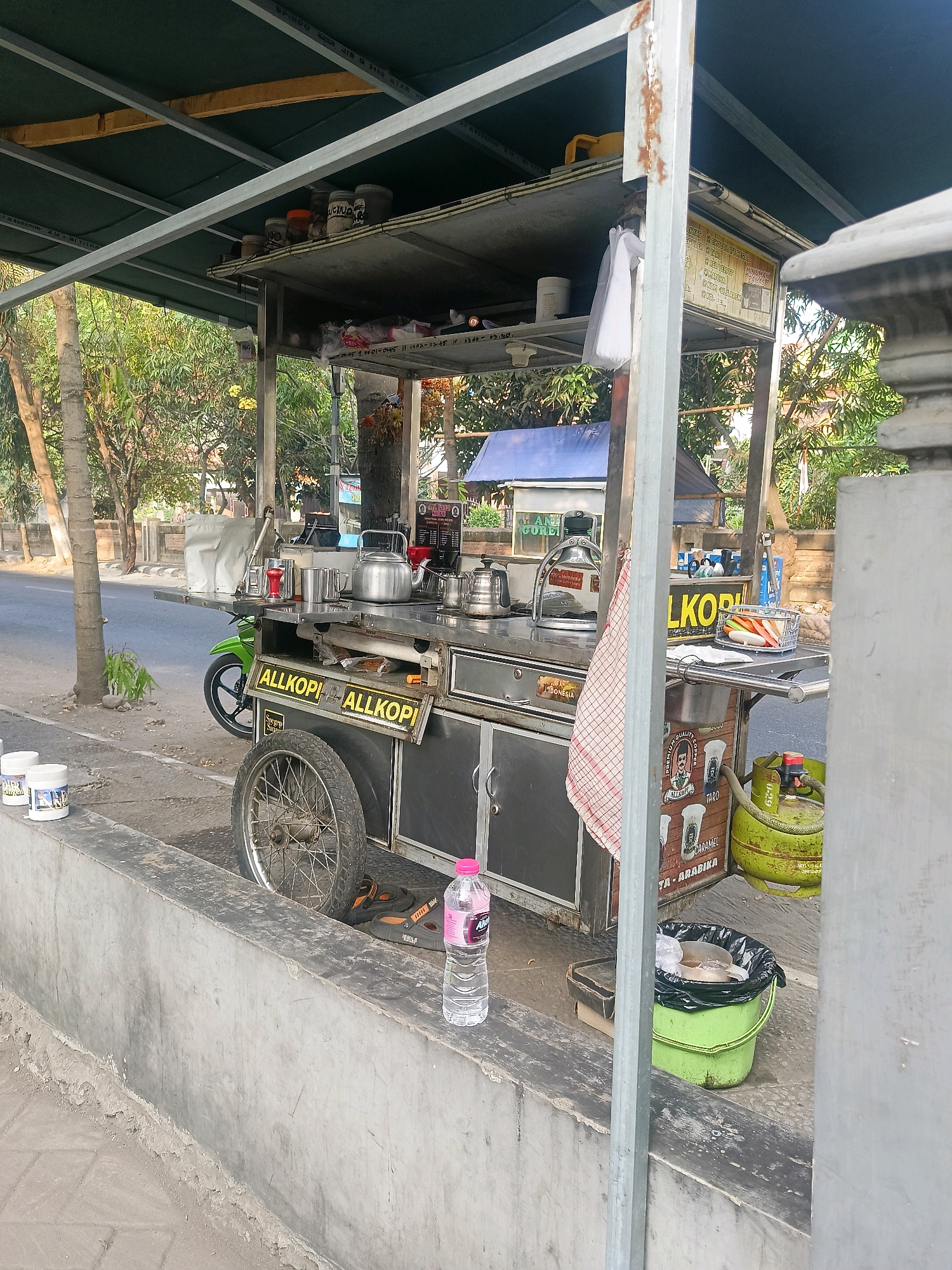 A coffee cart is parked on a street next to a concrete barrier. The cart is equipped with a canopy, shelves, cooking appliances, and various containers. A green propane tank is attached to the cart, and a motorcycle is parked behind it. Several cups and bottles are visible on the cart and on the barrier. A trash can with a black liner is also present. Trees and buildings are visible in the background. The scene is outdoors and appears to be during the daytime.