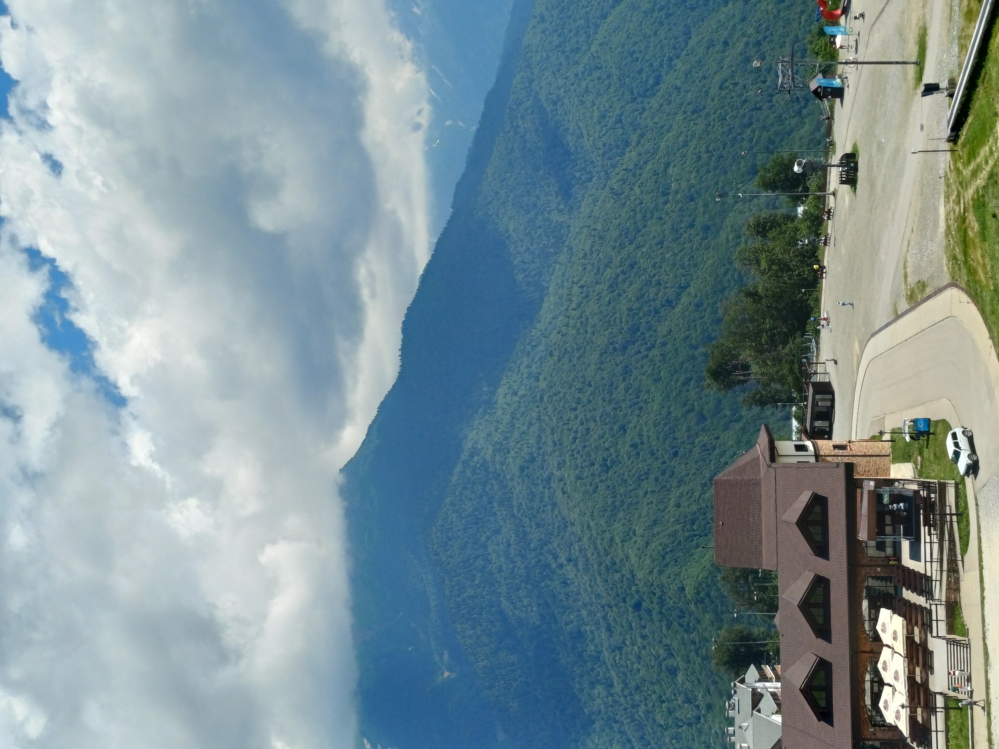 The image shows a mountainous landscape with a building complex in the foreground, likely a resort or a ski lodge in Estosadok, Russia. The sky is partly cloudy, with dramatic formations of white and grey clouds. Majestic, densely forested mountains dominate the background, with their peaks partially obscured by mist or low-hanging clouds. In the foreground, a building with a brown, multi-gabled roof and stone facade is visible. It has several outdoor seating areas with umbrellas. A paved road curves around the building, and a white car is parked nearby. Several people are scattered around the area, some walking and others appearing to be seated or gathered. A ski lift or similar structure is visible in the middle ground, along with a few lampposts and other infrastructure associated with a recreational area. The overall impression is one of a scenic mountain location during daylight.