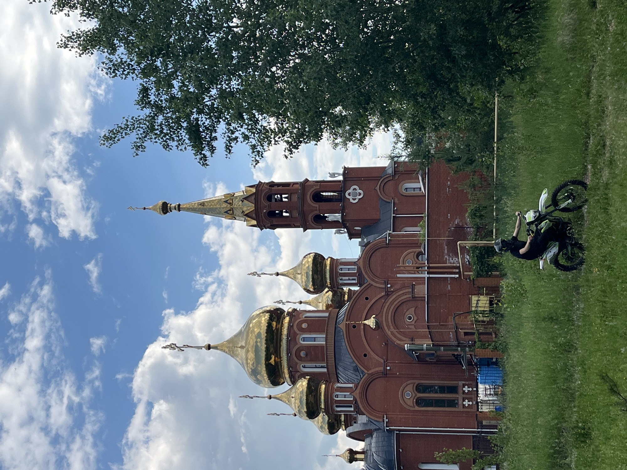 The image shows a red brick church with golden domes under a cloudy blue sky. In the foreground, a person wearing a black shirt, helmet, and protective gear is riding a dirt bike through a grassy field. The church appears to be a religious building, likely an Orthodox church, given the style of the domes and crosses. The surrounding environment is predominantly green, with grass and trees visible. The presence of the dirt bike rider suggests a recreational or possibly unauthorized activity in the vicinity of the church. This scene is located in the city of Chaikovsky, Russia.