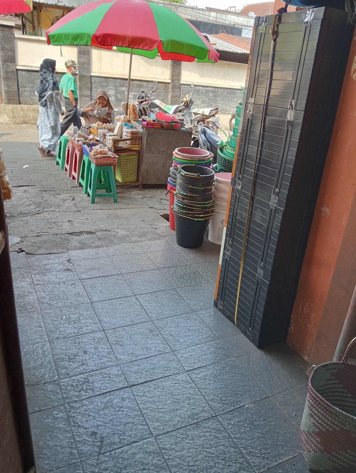 In Grabag, Indonesia, an outdoor scene depicts a market stall with a woman tending to it. She is seated behind a display of snacks arranged on small green and red stools and a simple table. Two individuals, a man wearing a green shirt and a cap, and a woman in a patterned dress and hijab, are walking away from the stall. A red and green umbrella is propped up over the stall. In the foreground, a stack of black buckets with yellow handles and various colored plastic containers are visible. To the right, a tall, black, folded structure leans against an orange wall. Further right, a woven basket rests on the tiled ground. The background shows buildings with corrugated roofs and a brick wall.