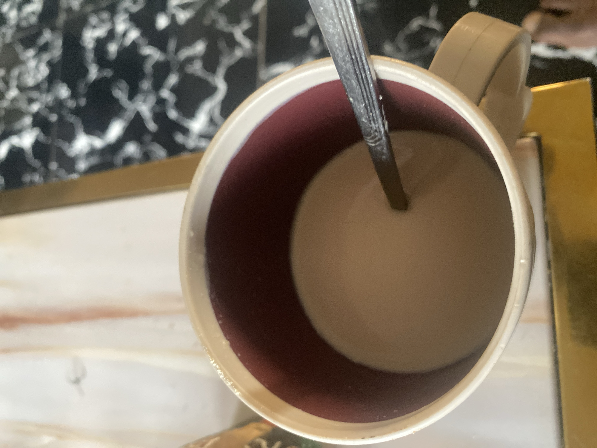 The image provides a high-angle, close-up view of a ceramic mug filled with a light-colored beverage, likely coffee or tea with milk. A metallic spoon, possibly stainless steel, is inserted into the liquid. The mug has an off-white or light beige exterior and a prominent dark reddish-brown interior lining around the rim, which contrasts with the lighter, creamy color of the drink inside. The handle of the mug, also off-white, is partially visible on the right side.
The mug is situated on a multi-textured surface. To the upper left, a dark background with striking white, almost marbled or speckled, patterns is visible. Beneath and to the left of the mug, there is a lighter, subtle surface, possibly a light wood or stone pattern, with faint, lighter brown striations. On the far right, a shiny, golden-colored object or surface, perhaps a tray or part of a table, is partially captured.
The scene suggests an indoor setting, such as a kitchen or dining area, in Abuja, Nigeria, implying a moment of rest or refreshment. There are no people visible, and no discernible text. The time of day and weather conditions cannot be determined from the image.