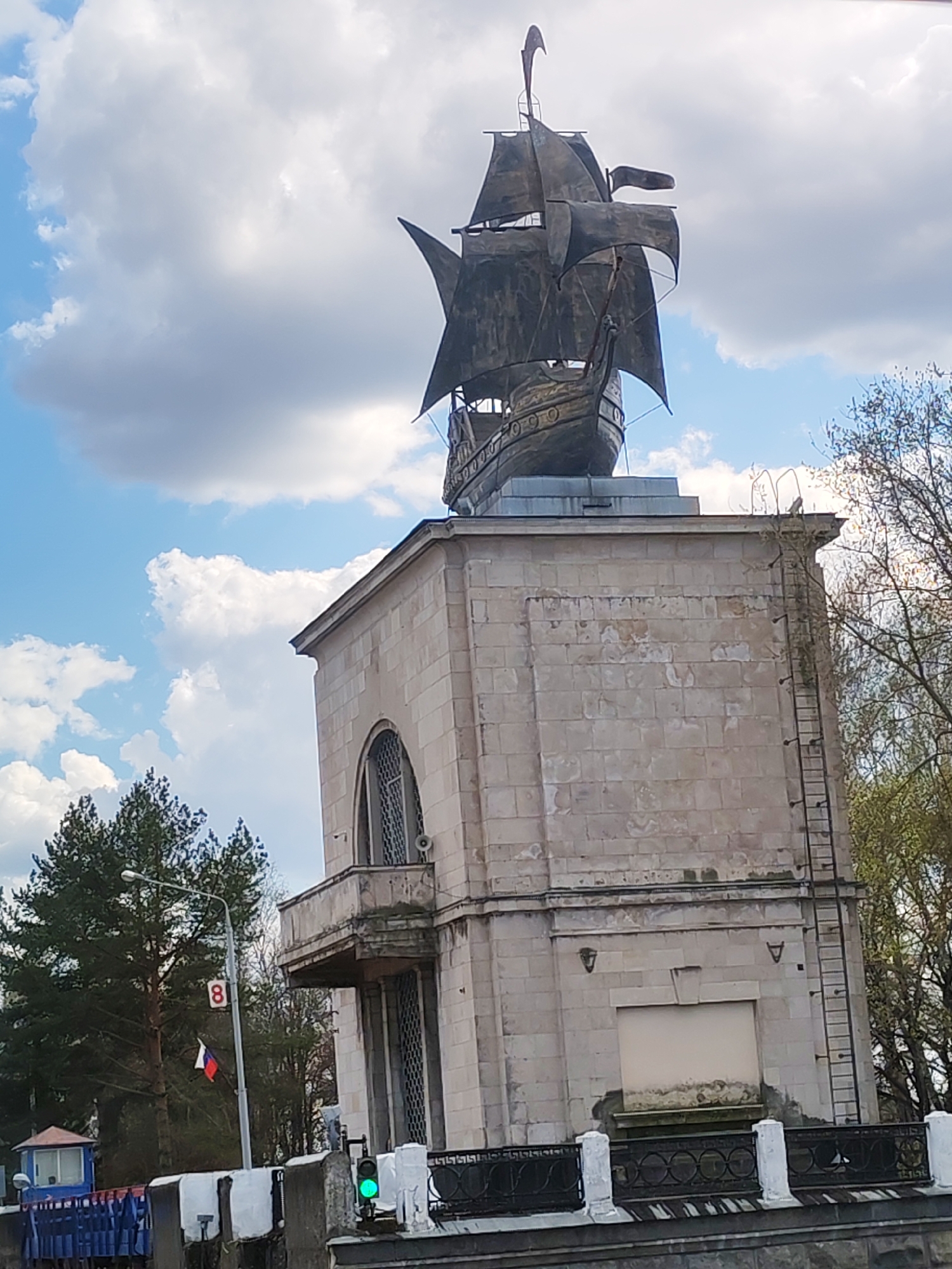 The image captures a statue of a ship perched atop a stone structure, likely a monument, set against a partly cloudy sky. The statue is detailed and depicts a sailing ship with multiple masts and sails. The stone structure beneath is rectangular with architectural details like arched windows and a ladder extending up its side. The structure's surface shows signs of weathering.
In the foreground, there is a black iron railing and a section of what appears to be a blue fence. Beyond this, a street scene includes a street lamp with a sign marked "8" and a Russian flag. A small blue guard house is also visible. To the sides of the monument, trees suggest an urban or suburban setting.
The location is likely Yakhroma, Russia, based on the monument's style, architectural elements, and cultural symbols present in the photograph.