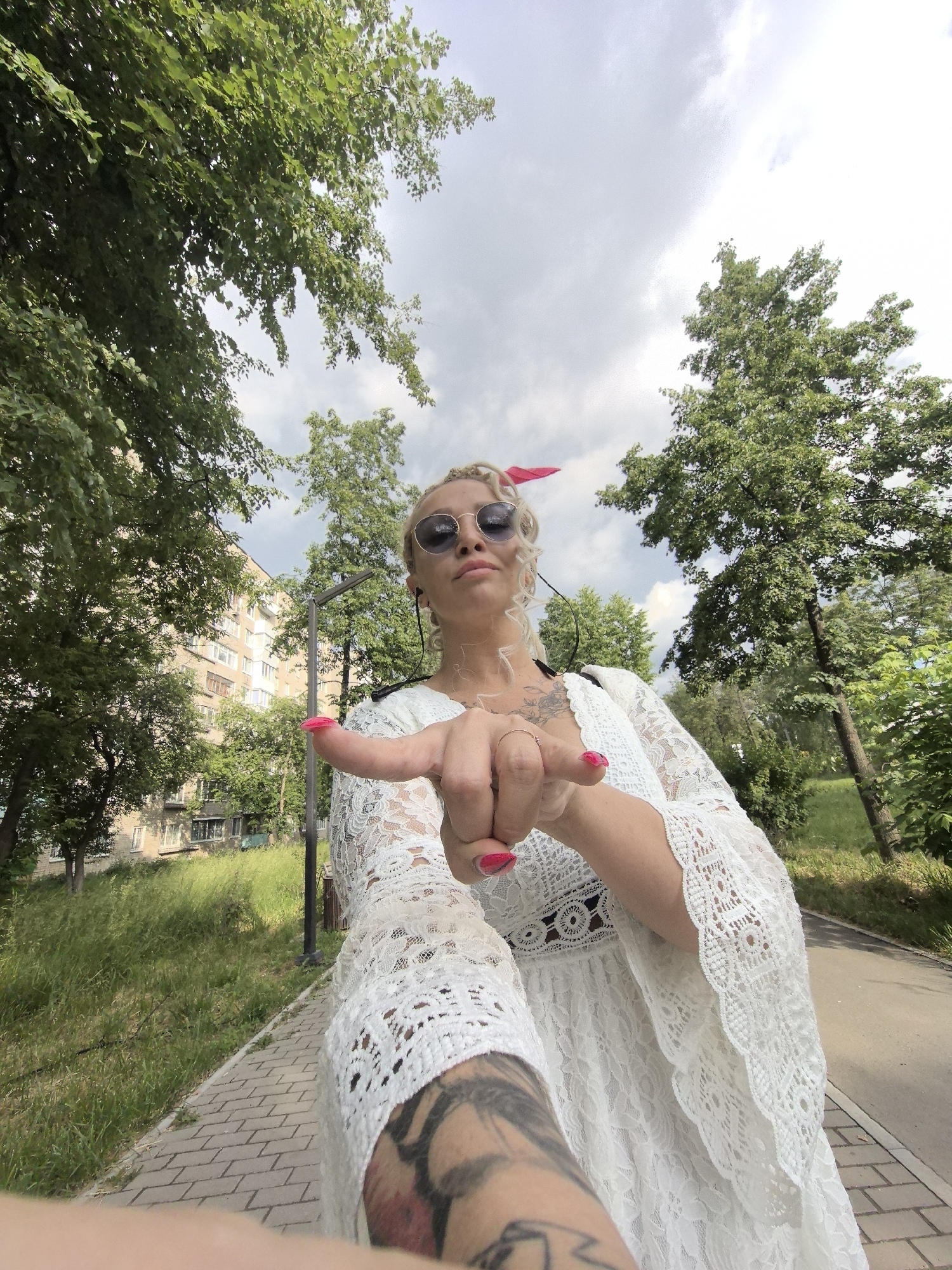 A woman with blonde braided hair, wearing sunglasses and a white lace dress, points her finger towards the camera. She has bright pink nail polish. The background shows a street with trees, a building, and a cloudy sky. The scene is outdoors in Chaikovsky, Russia.