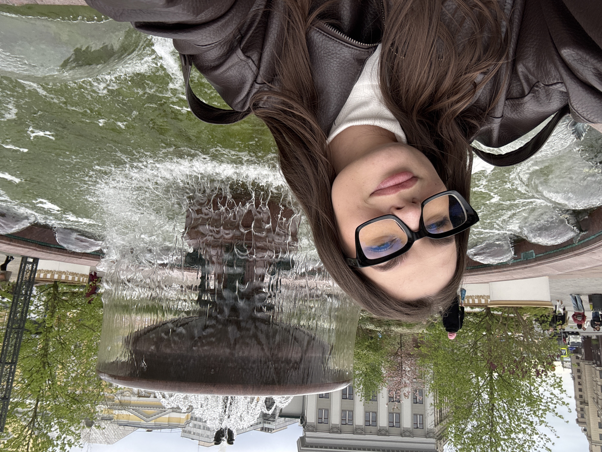 A young woman with long brown hair and wearing glasses, a white turtleneck, and a brown leather jacket, is taking a selfie. She is standing in front of a large fountain. The fountain appears to be made of dark stone and has multiple tiers, with water cascading down. In the background, buildings are visible, suggesting an urban setting. The scene is in Moscow, Russia. Other people are present in the background, walking around the park or square where the fountain is located. Trees are also visible, indicating it may be a park or garden area.