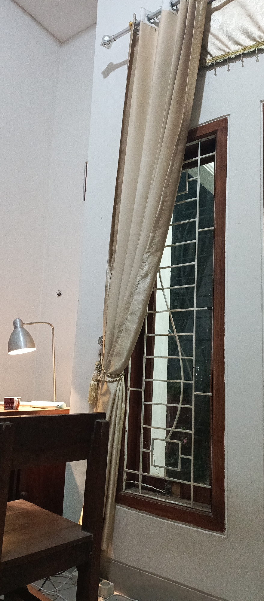 The image depicts an indoor scene with a window, a desk, a chair, and a lamp. The window is covered by a beige curtain, and a metal grille is visible on the glass. On the desk, there is a mug and a lamp with a silver shade. A dark wooden chair is partially visible in the foreground. The wall is painted white, and there is a small object resembling a fly on the wall. The scene suggests a study or workspace. The location is Jatisari, Indonesia, which is a region known for its agricultural activities and rural landscapes.