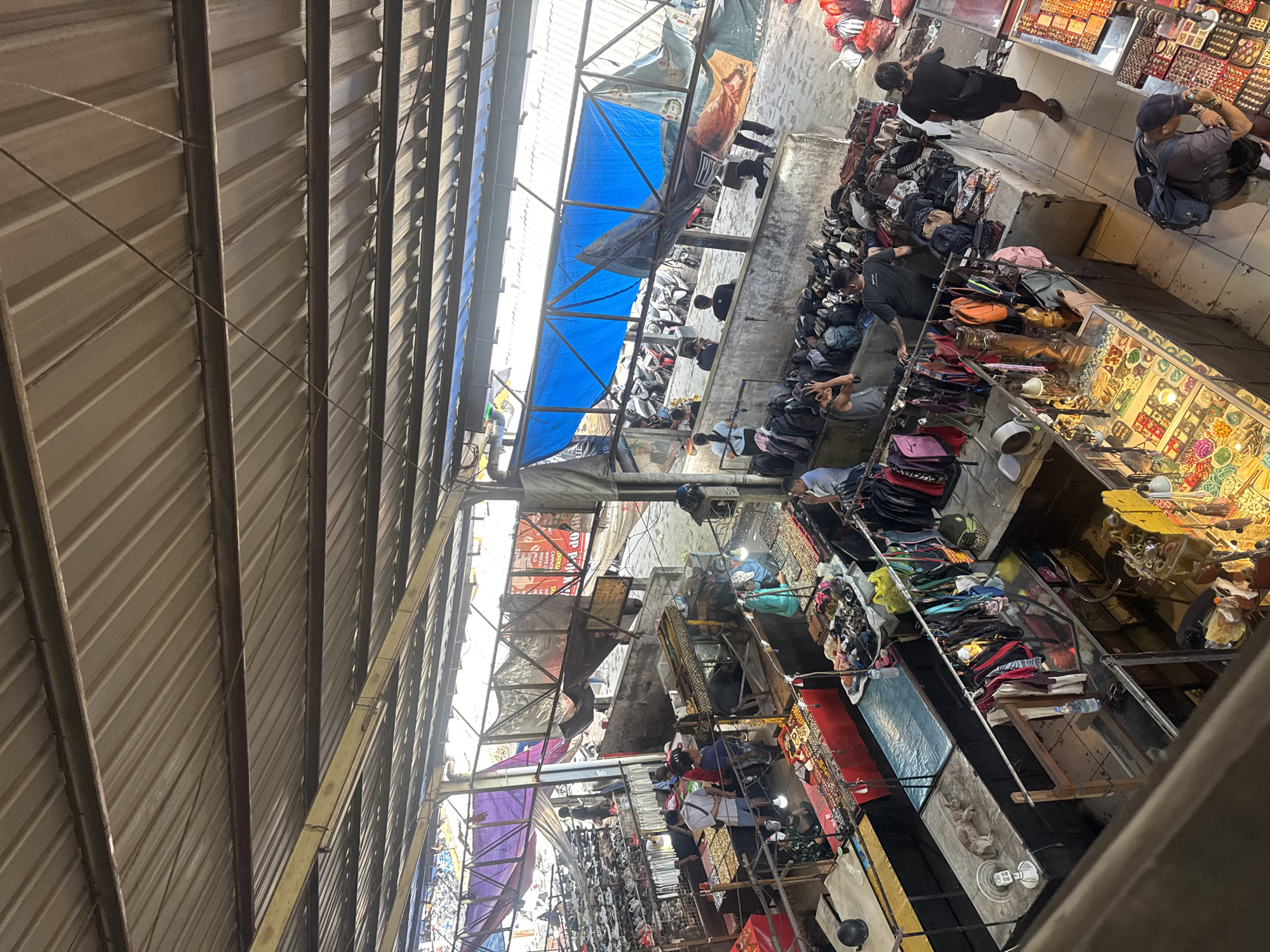This is an elevated, full shot of a busy outdoor market in Denpasar, Indonesia. The market is covered by a corrugated metal roof supported by beams. Stalls are set up with various goods for sale, including clothing, bags, and jewelry. People are seen browsing and interacting at the stalls. The ground is paved and appears to be a mix of concrete and stone. In the background, several motorcycles are parked. The overall atmosphere is one of commerce and activity.