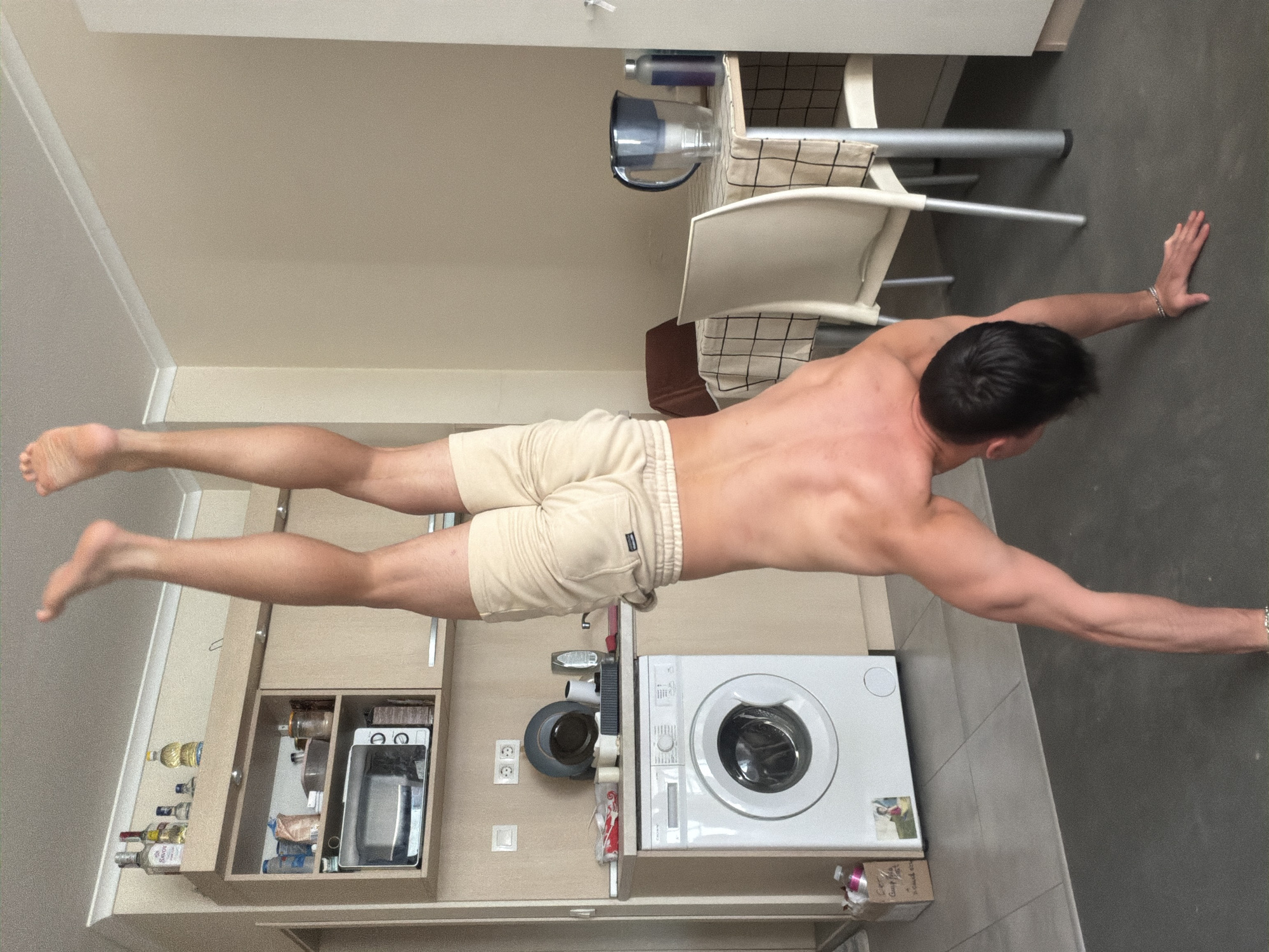 A man is performing a human flag pose in a kitchen. His body is horizontal, supported by his hands against a wall. He is wearing beige shorts. A washing machine, kitchen cabinets, and a chair are visible in the background. The location is a kitchen within a residence, possibly in Sofia, Bulgaria, based on architectural style.