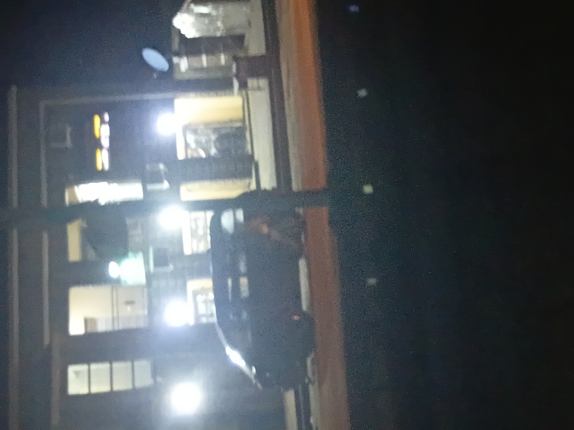 The image shows a dark, grainy scene at night, likely in Jalingo, Nigeria. In the foreground, a dark-colored van or SUV is parked on a street, its shape slightly obscured by the low light. Behind the vehicle, a building with multiple windows is visible. Some windows are lit from within, casting bright white and yellow glows. A prominent vertical pole, possibly a street light or signpost, stands in front of the building and partially obscures the van. The overall atmosphere is dimly lit and somewhat hazy.