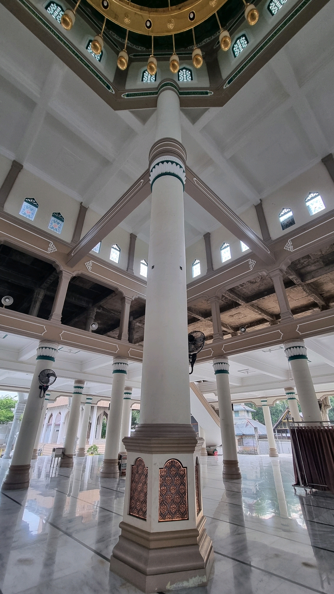 This is a low-angle, wide shot of the interior of a mosque in Lhokseumawe, Indonesia. The central focus is a massive, octagonal pillar with ornate bronze-colored panels near its base. Above, the pillar transitions to a cylindrical shaft, culminating in a decorative capital with green and white bands. The pillar supports an intricate ceiling structure featuring a large, gold-colored chandelier with multiple light fixtures.
Surrounding the central pillar, rows of smaller, similar pillars extend into the background, supporting an upper level with arched openings and exposed structural elements. Smaller windows with decorative grilles are visible along the upper walls. The floor is polished marble with subtle gray veining and dark lines. Several black oscillating fans are mounted on some of the pillars. In the background, glimpses of the outdoors show greenery and building structures. The overall atmosphere is one of grandeur and architectural detail.