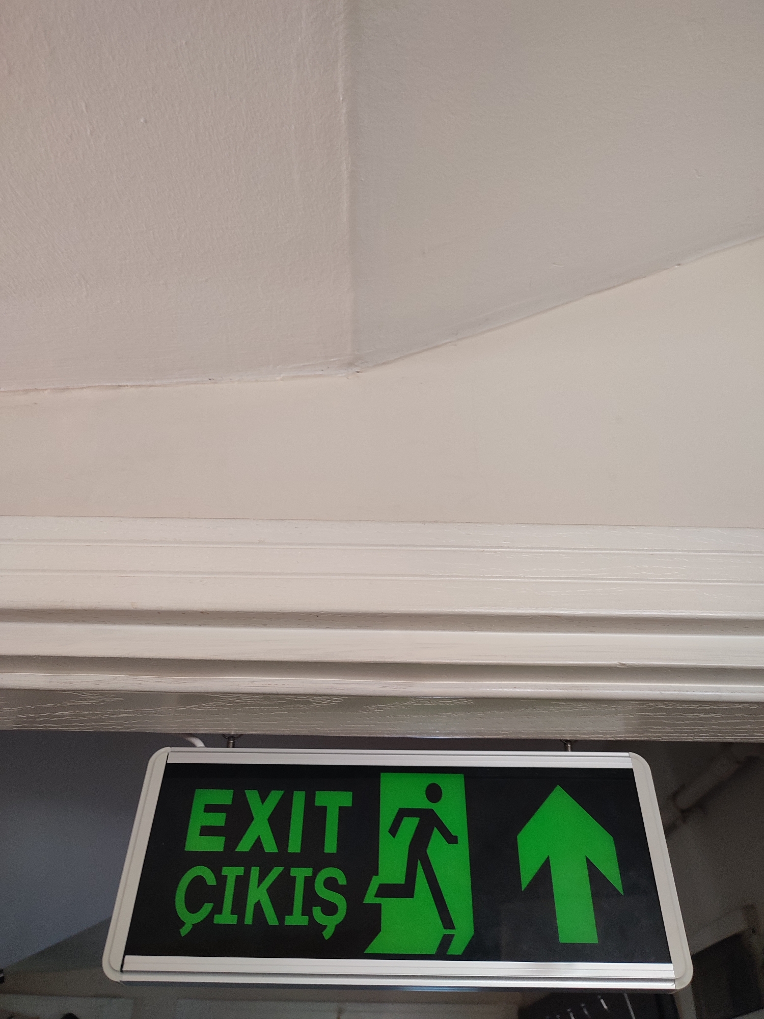 A green and black illuminated exit sign is visible in the foreground, mounted above a doorway. The sign displays the word "EXIT" and "ÇIKIŞ" in green text, along with a running man icon and an upward-pointing arrow, all in green against a black background. The sign is attached to a white ceiling with a detailed architectural molding below it. The ceiling transitions from a lighter shade on the left to a slightly darker shade on the right, with a clear seam running diagonally. The environment appears to be indoors, possibly a hallway or entryway, in Üsküdar, Turkey.