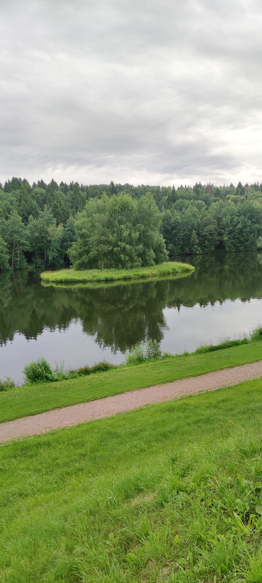 The image captures a serene natural landscape at Росинка Кп, Russia. The scene is dominated by a tranquil lake reflecting the lush greenery of the surrounding forest. In the middle of the lake, a small, verdant island covered in grass and trees adds a focal point. The background is a dense forest composed of various shades of green trees, creating a rich texture. Above, the sky is overcast with soft, diffused light, suggesting it might be a cloudy day or perhaps early morning or late evening. A dirt path runs diagonally across the lower part of the image, bordered by vibrant green grass on both sides, indicating a well-maintained park or recreational area. The overall atmosphere is peaceful and idyllic, with no people or specific objects other than the natural elements and the path. The absence of direct sunlight and the cloudy sky contribute to a calm, almost muted ambiance.